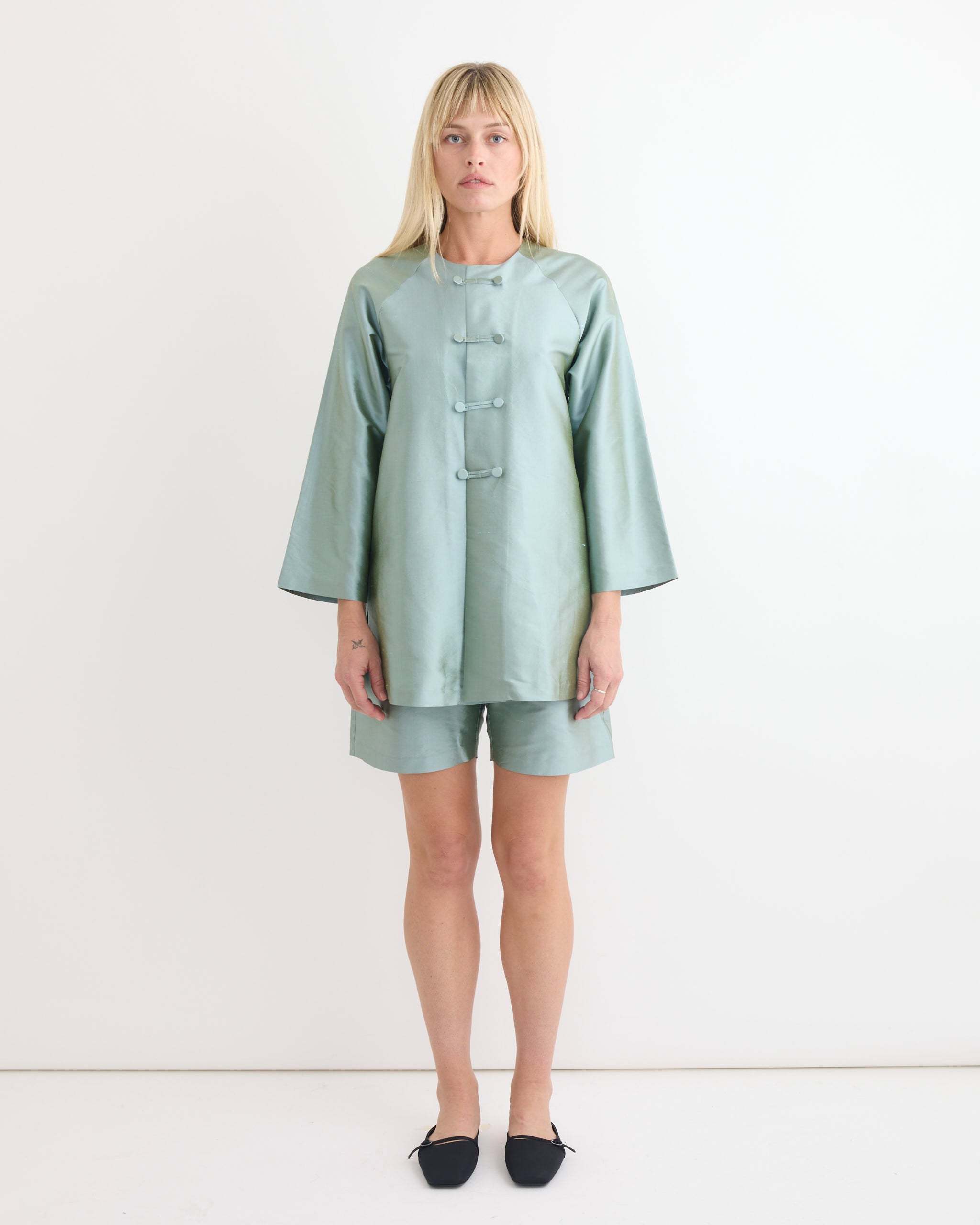 A woman with straight blonde hair and bangs stands against a white wall, wearing the Chelsea Mak Otto Short in Mint Taffeta, size XS, paired with a matching light green satin tunic and black pointed-toe flats.