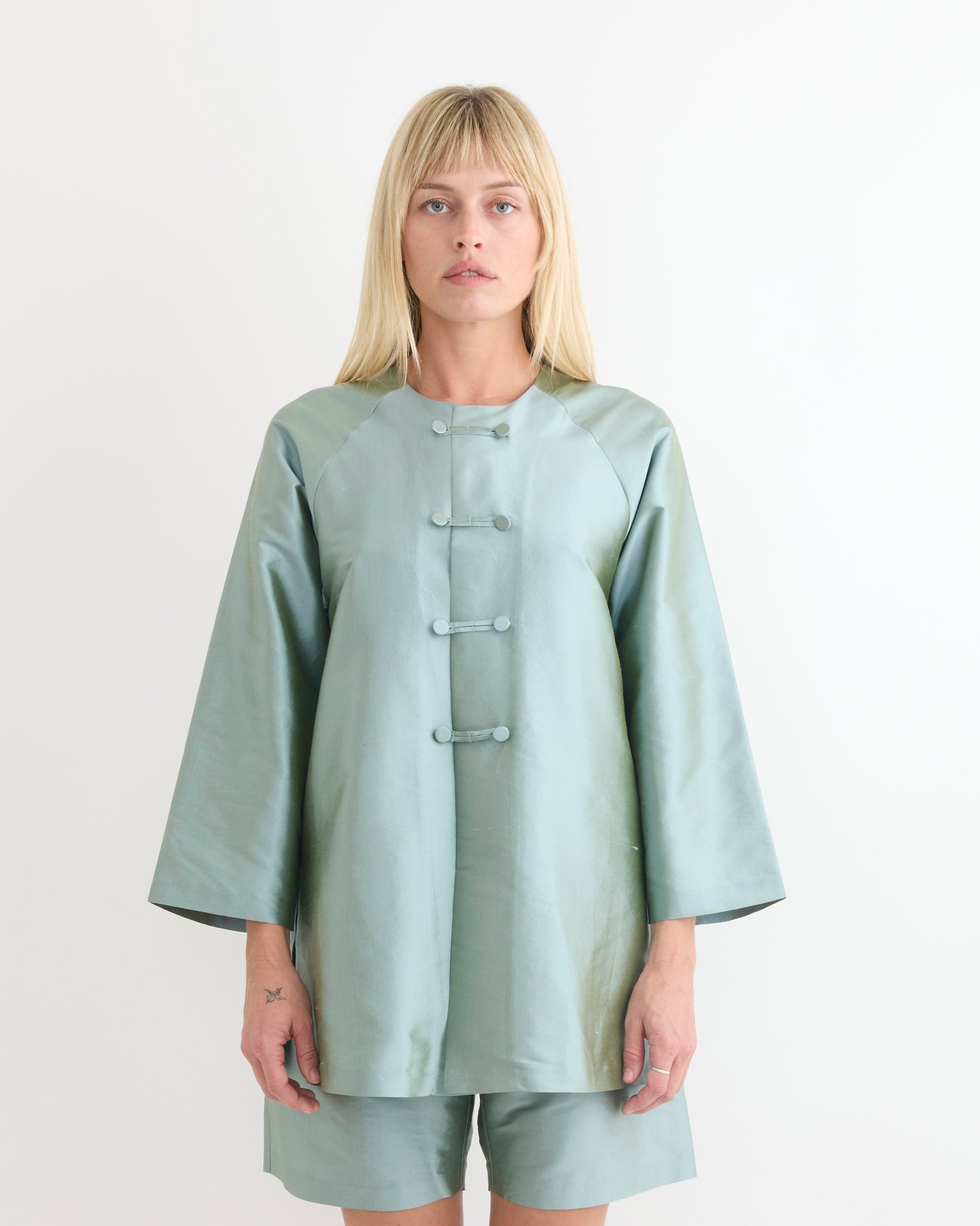 A person with straight blond hair wears the Chelsea Mak Luc Jacket in Mint Taffeta, featuring frog button closures, standing against a plain white background. For fit or sizing questions, our online chat is available to help.