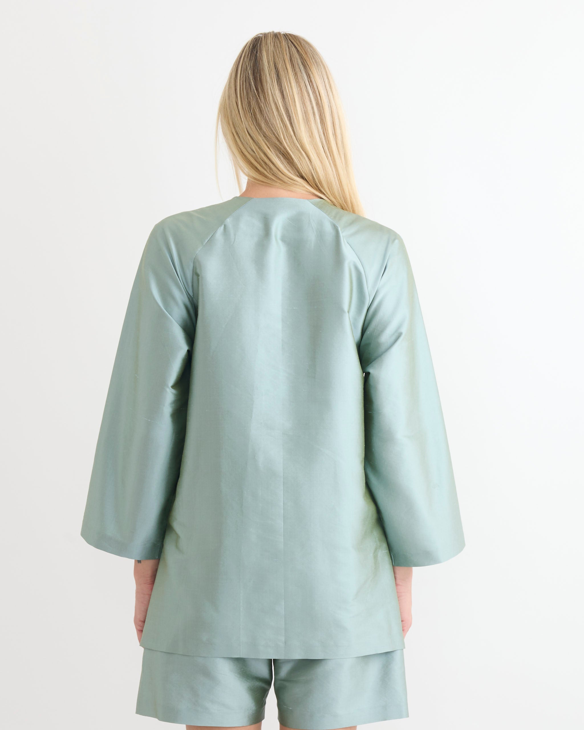 A person with long blonde hair faces away, wearing the Chelsea Mak Luc Jacket in Mint Taffeta against a plain white background. The relaxed fit makes it perfect for easy sizing when shopping online.