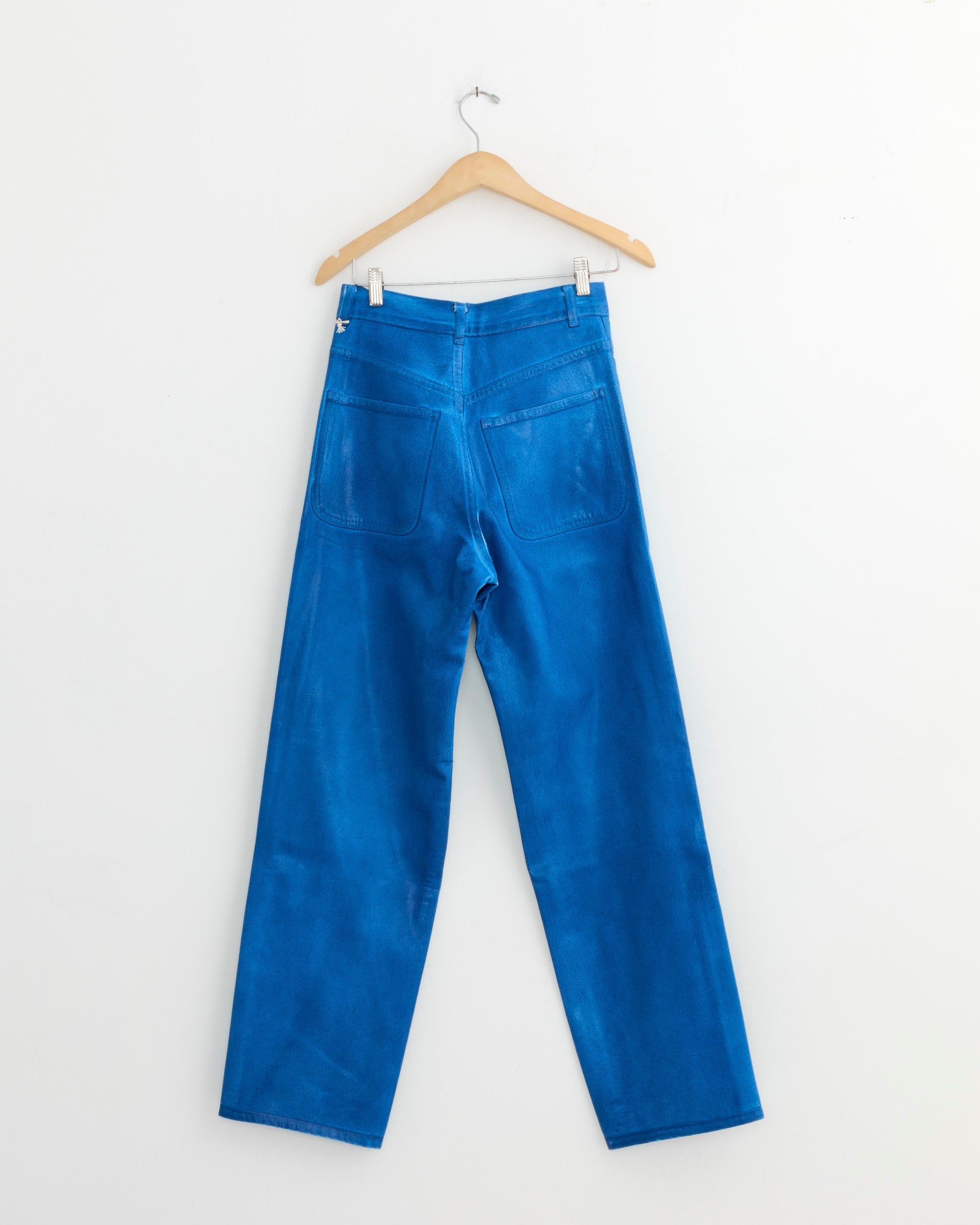 The 225 in Electric Blue Lacquer by Jesse Kamm, regenerative Japanese denim jeans, hang on a wooden hanger against a plain white wall, displayed from the back to showcase their two rear pockets.