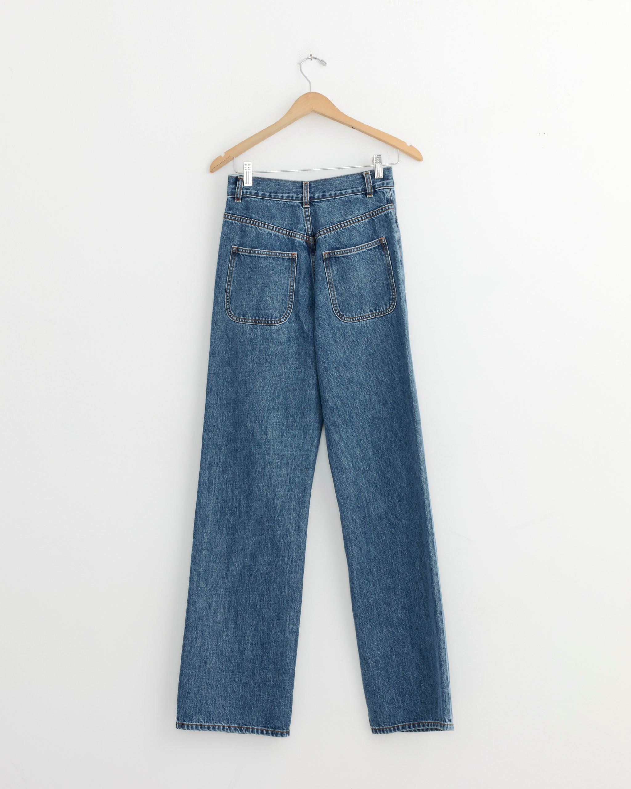 The Jesse Kamm Alta Jean in Cowboy Blue hangs on a wooden hanger against a plain white wall, displayed from the back with visible pockets and straight legs.