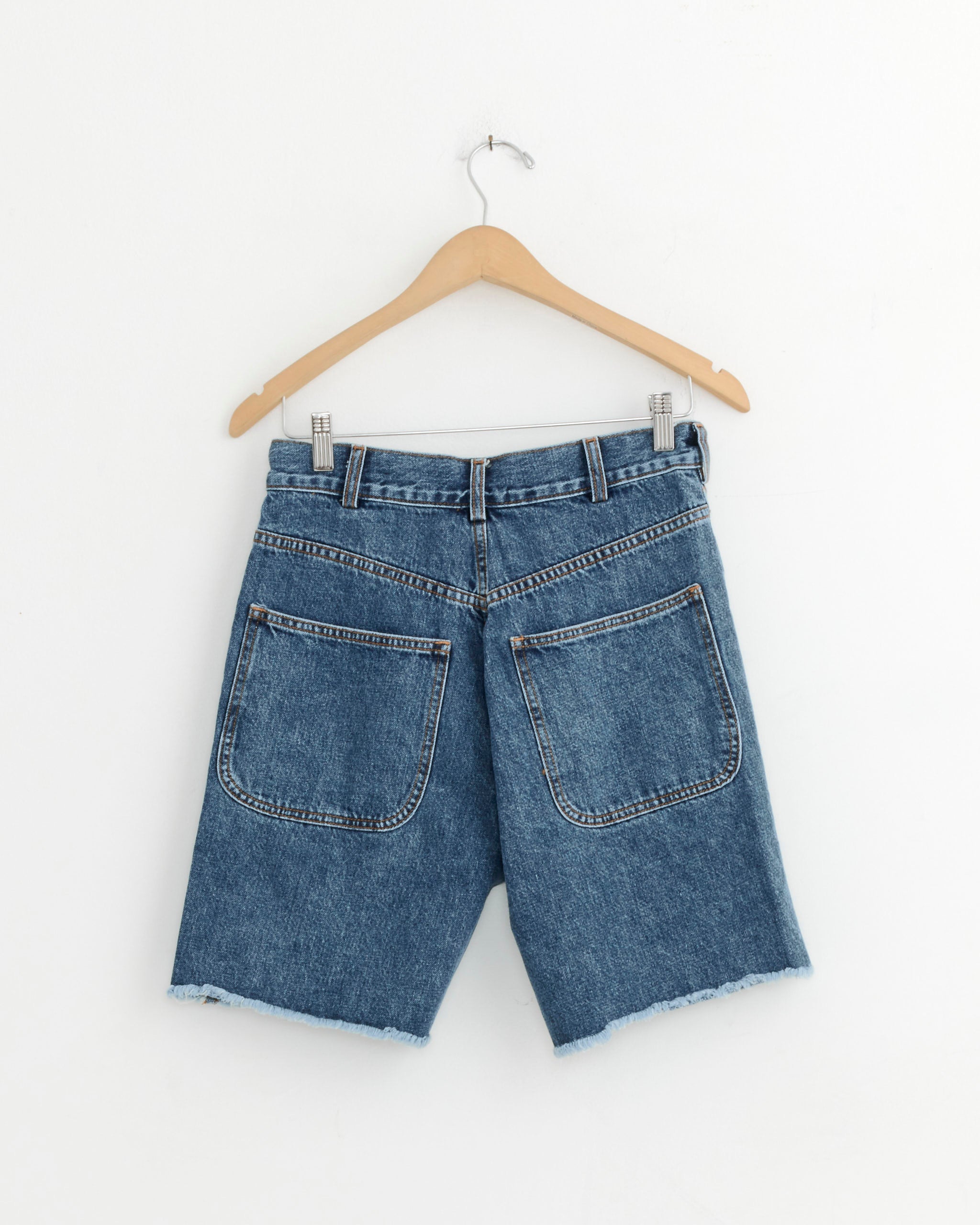 The Lake Short in Cowboy Blye by Jesse Kamm, made from regenerative Japanese cotton with frayed hems, is shown from the back on a wooden hanger against a plain white background.