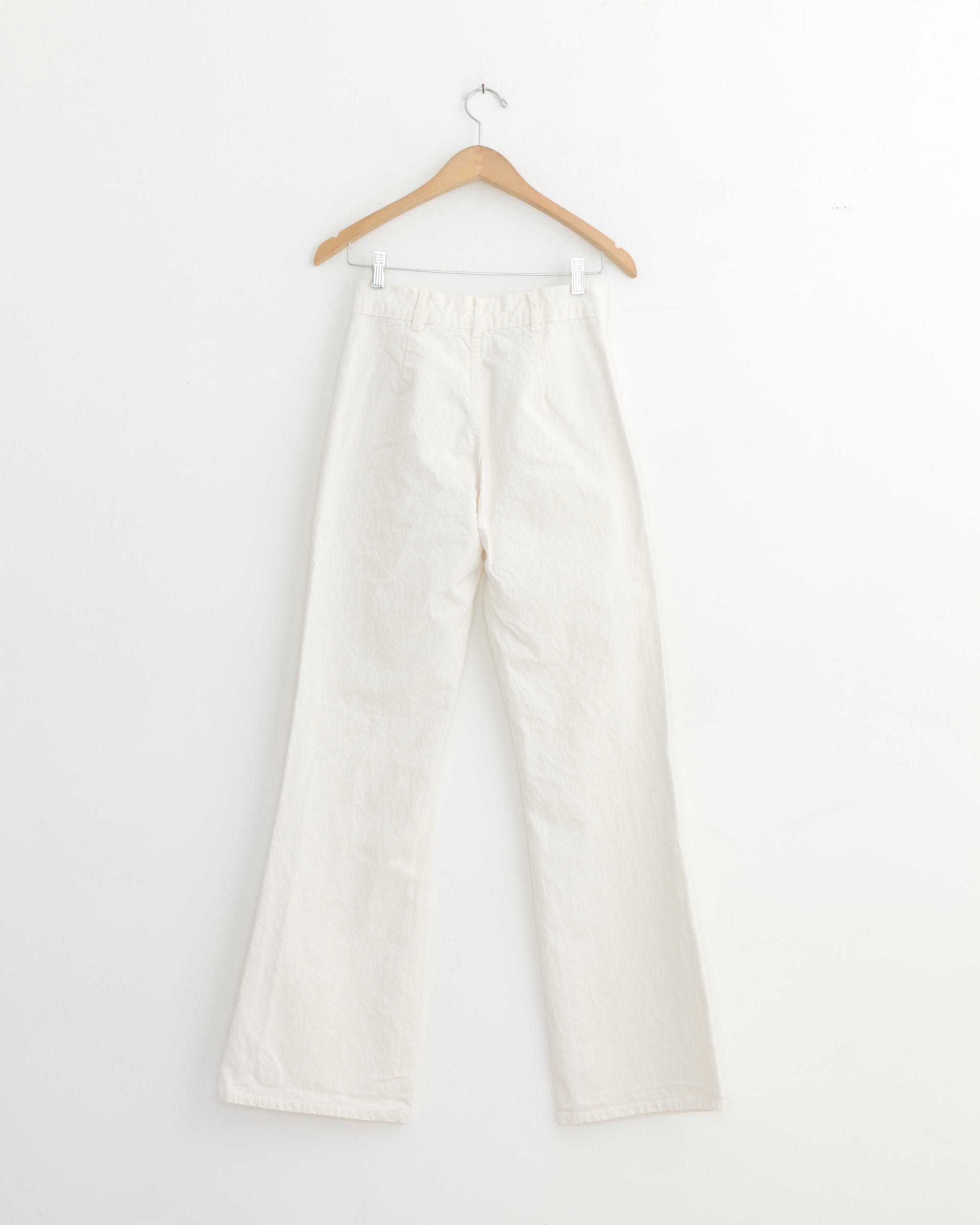A pair of Jesse Kamm Les Deux Pant in Salt White, crafted from organic cotton canvas, hangs on a wooden hanger against a plain white wall.