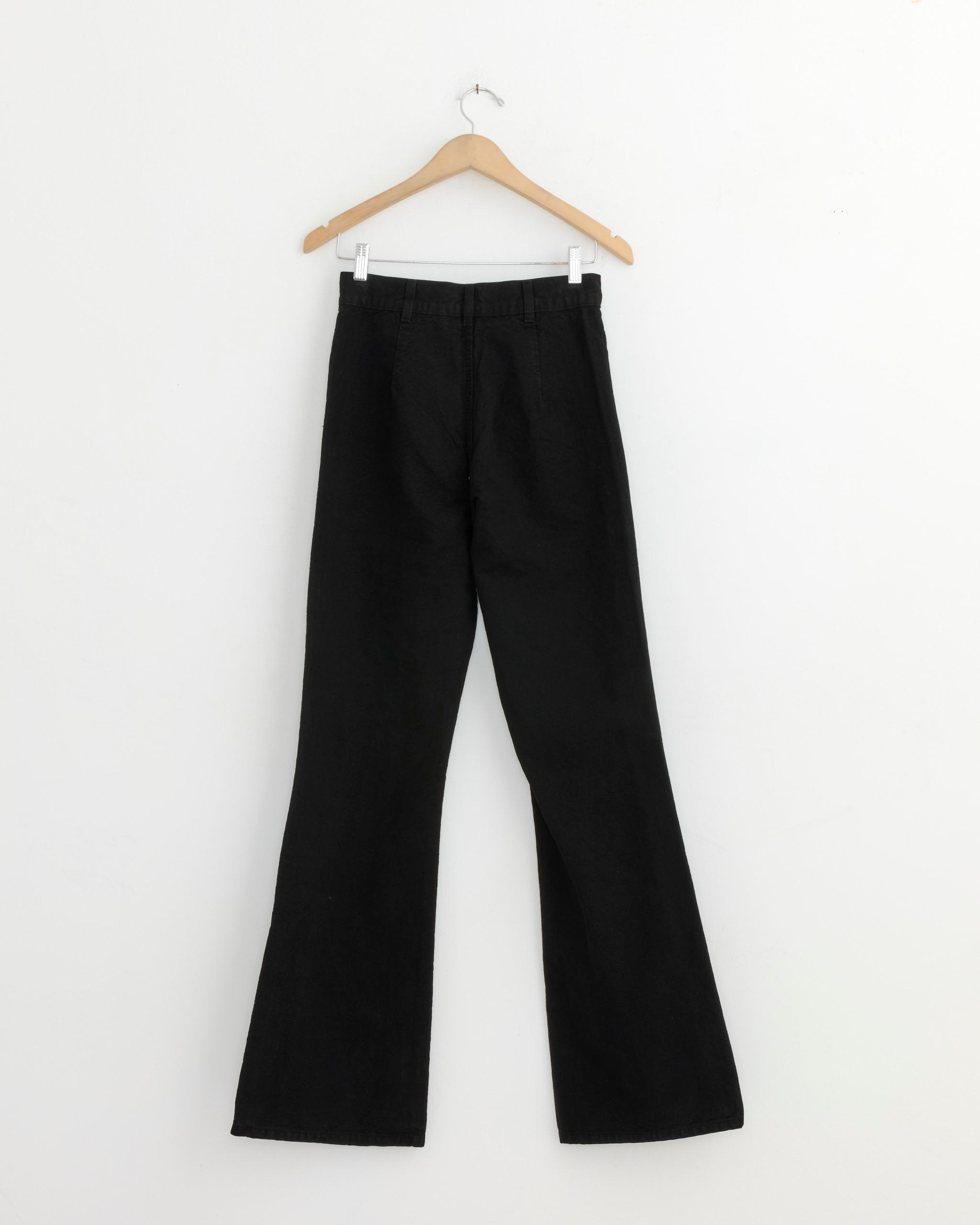The Jesse Kamm Les Deux Pant in Black, a high-waisted wide-leg style, hangs on a wooden hanger against a plain white wall.