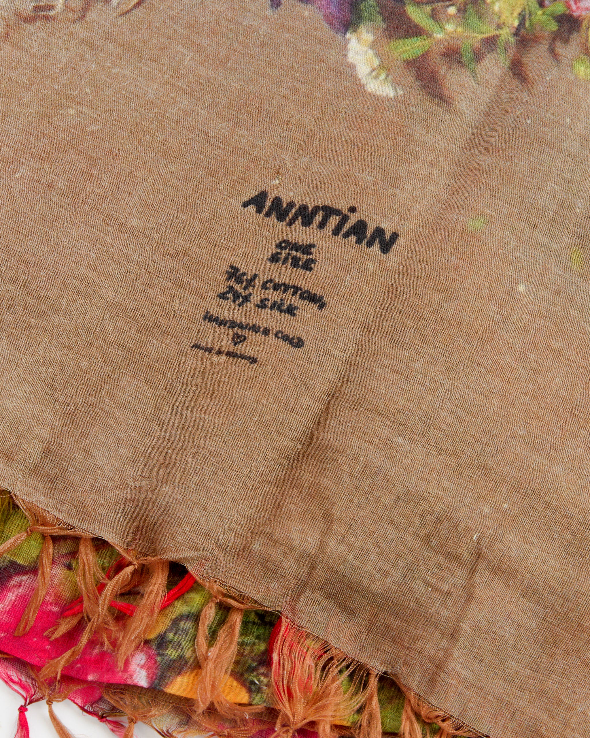A close-up of Scarf in Print X by Anntian features a floral print in brown with colorful patterns and orange fringe. The one size scarf is 74% cotton, 26% silk—elegant and made in Germany. Hand wash cold.