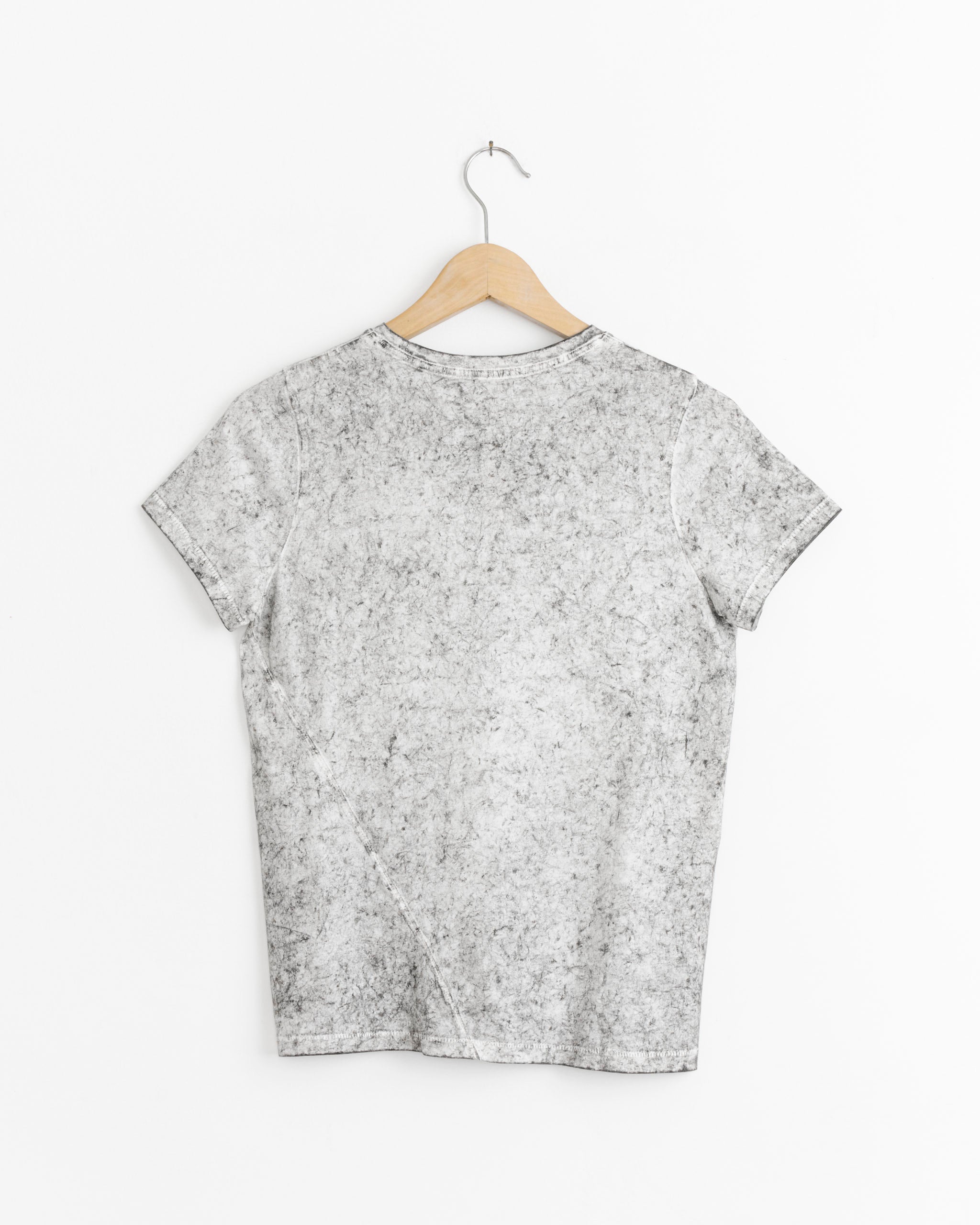 The Lauren Manoogian Bias Baby Tee in Rock Crackle, a slim-fit crew neck with a light gray crackled texture, hangs on a wooden hanger against a white wall and is displayed from the back.