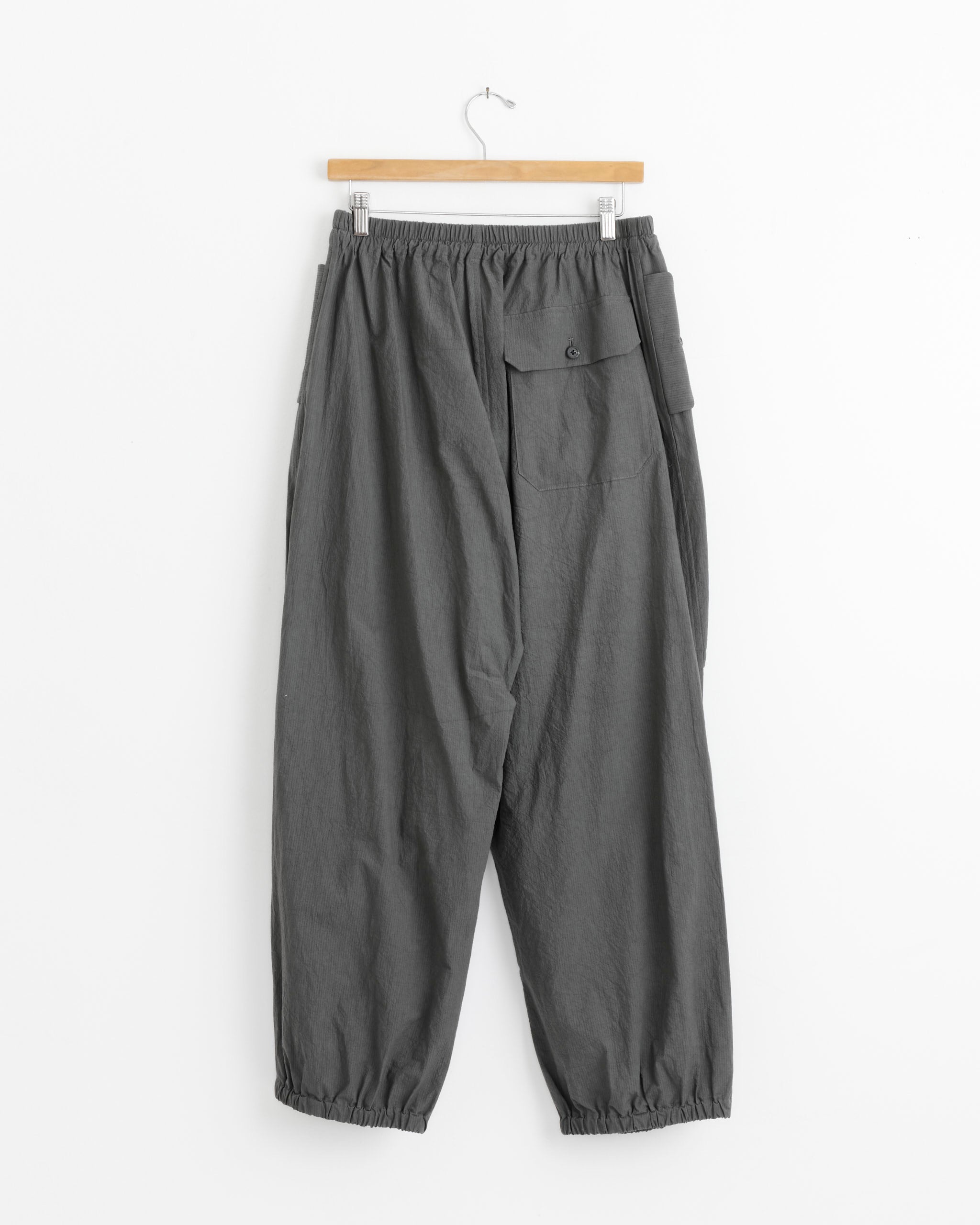 The Lauren Manoogian Ripstop Pant in Coal is a loose-fitting cotton style with an elastic waistband and cuffs, plus a right-side flap pocket, displayed on a wooden hanger against a white background.