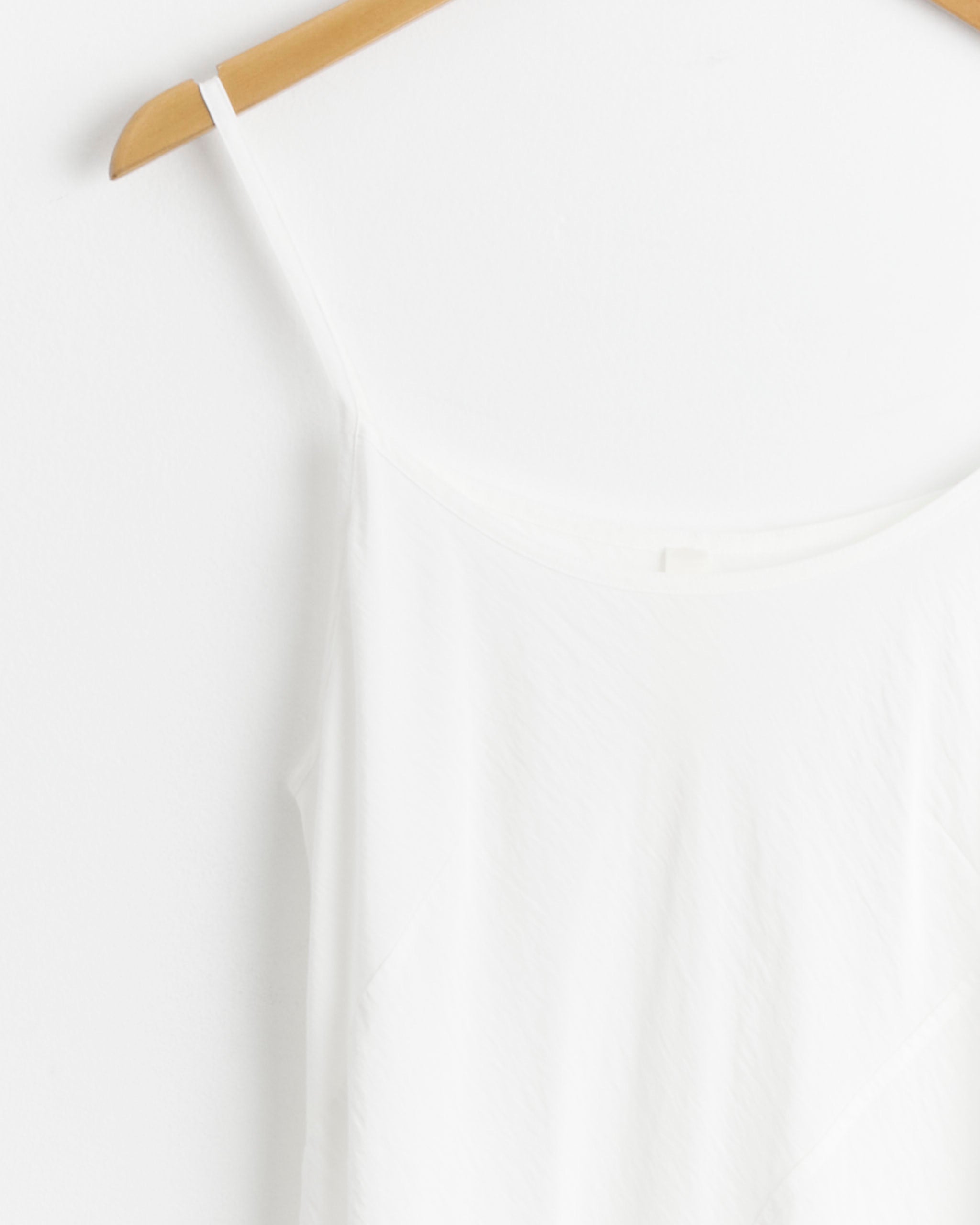 A close-up of the Lauren Manoogian Bias Slip in White, a summer slip dress with thin straps, hanging on a wooden hanger against a white background.