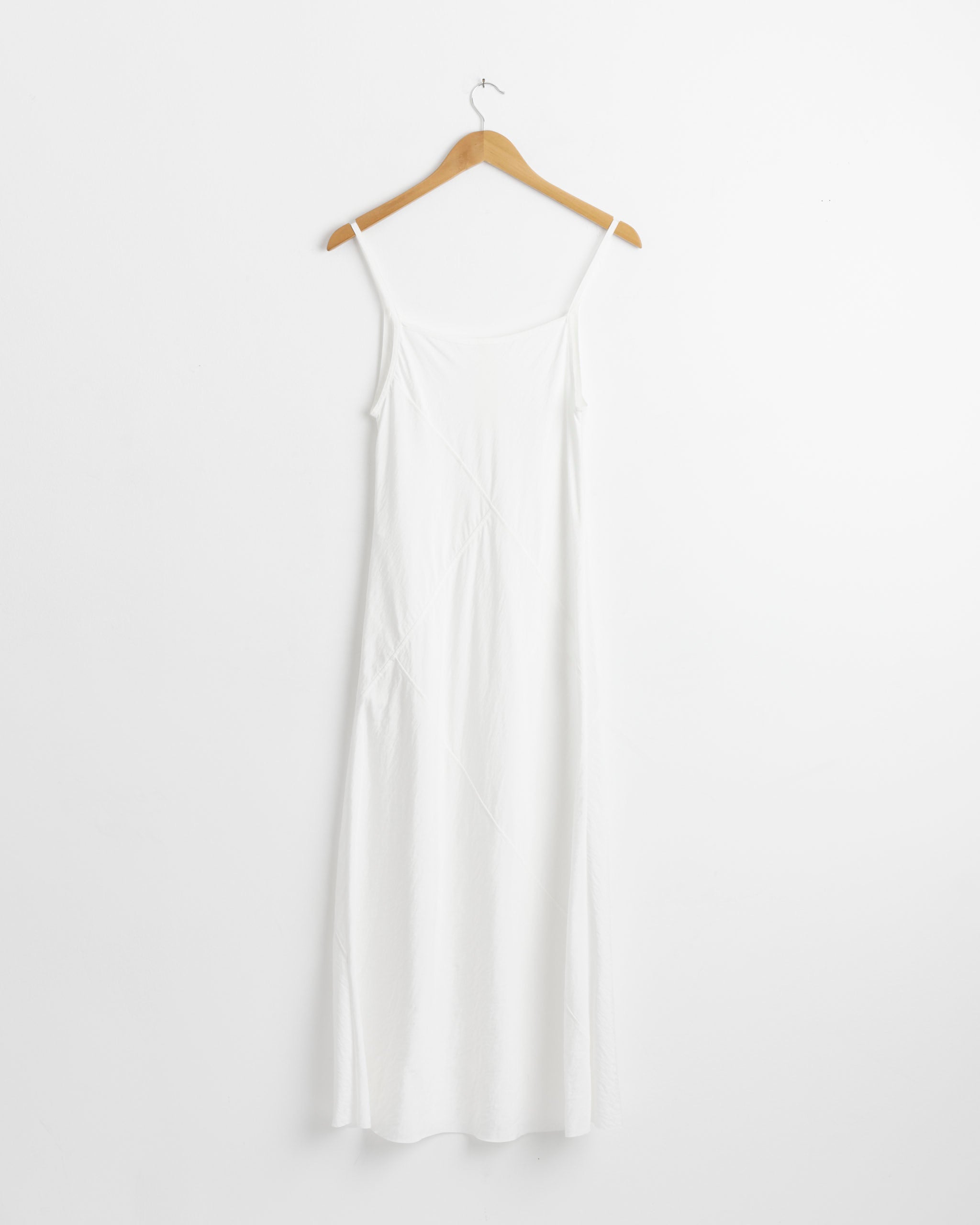 The Lauren Manoogian Bias Slip in White, a minimalist summer slip dress with thin straps, hangs on a wooden hanger against a plain white background.