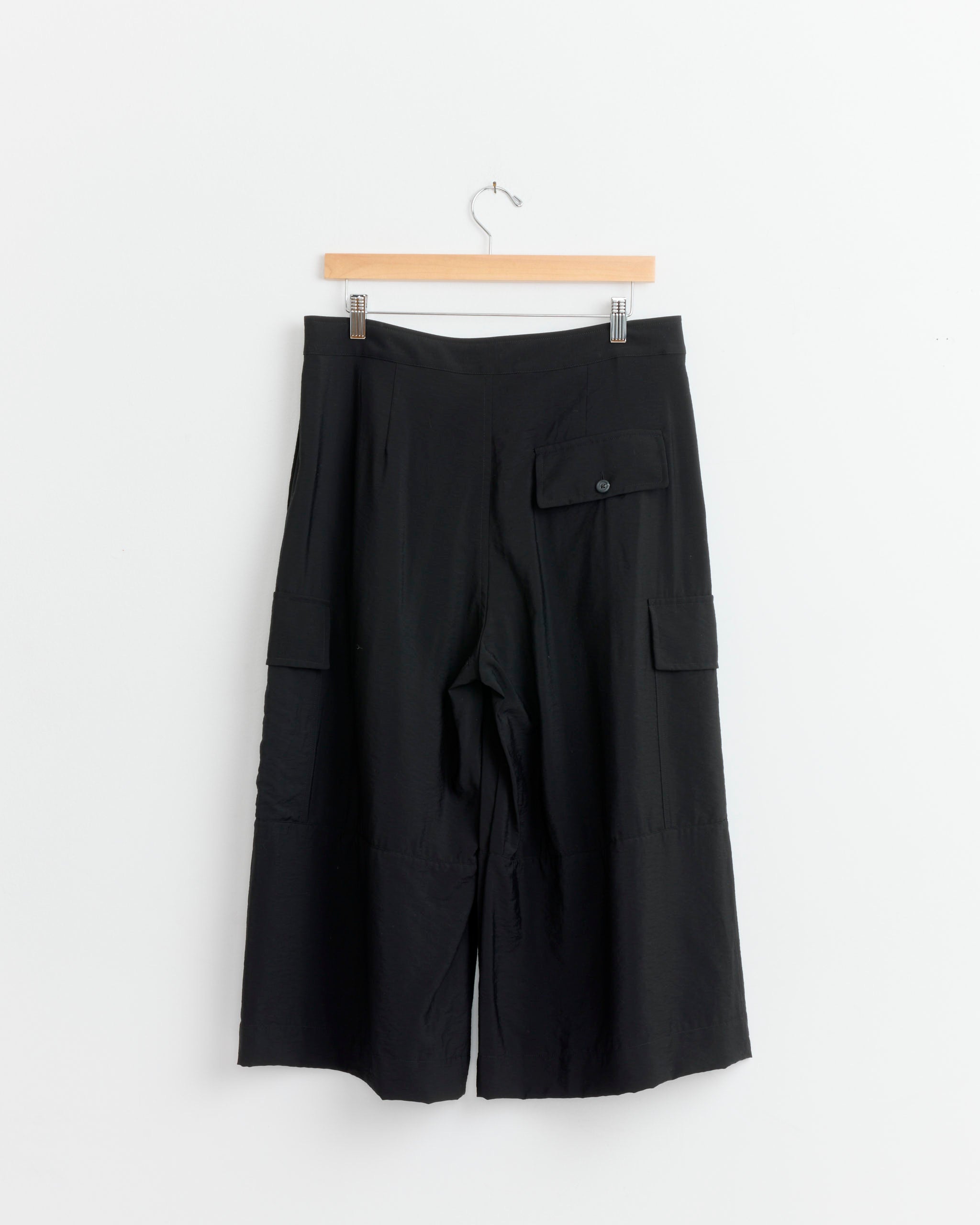 The Lauren Manoogian Cargo Pant in Black, featuring a relaxed utilitarian fit with large pockets, hangs on a wooden hanger against a plain white background.