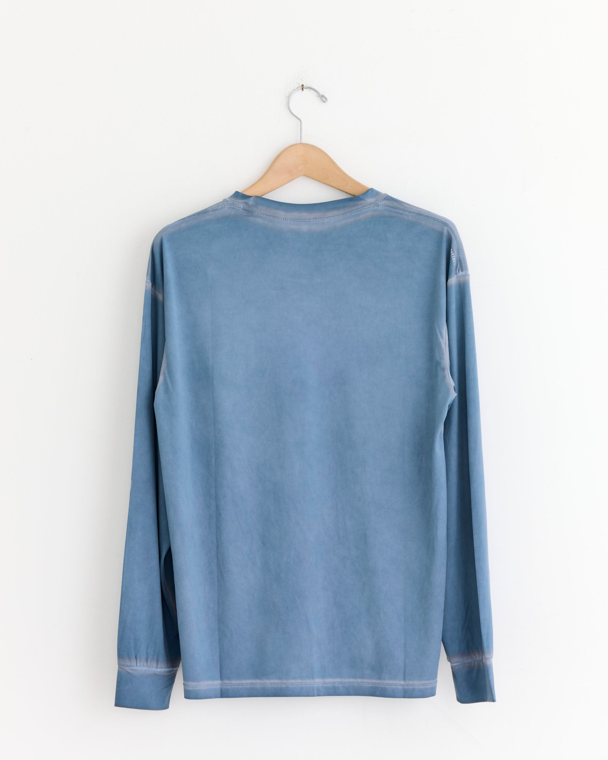 A plain, long-sleeve AuraLite™ Long Tee in Pigment Slate by Satisfy Running hangs on a wooden hanger against a white wall, shown from the back—made from recycled micro jersey for a lightweight running shirt feel.