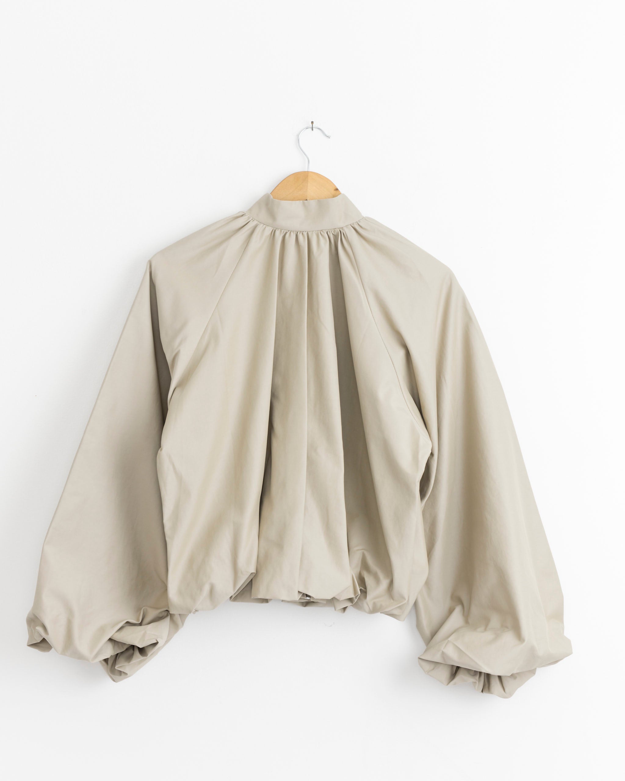 The Beaufille Tadao Jacket in Beige features sculptural balloon sleeves and a gathered back, showcased on a wooden hanger against a white wall.