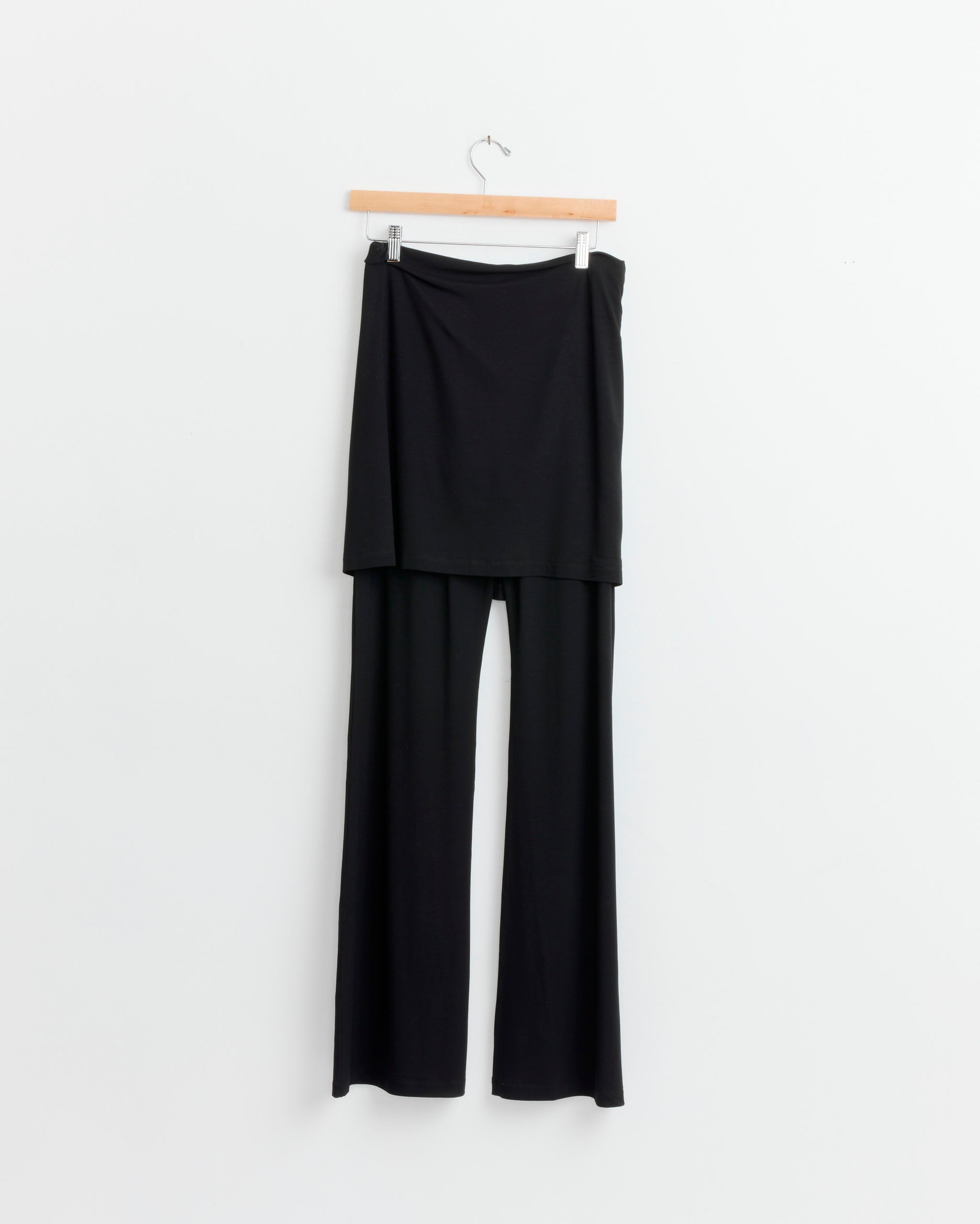 The Beaufille Waist Tie Pant in Black features flared legs and a skirt-like overlay, crafted from lightweight stretch fabric, displayed on a wooden hanger against a white background.