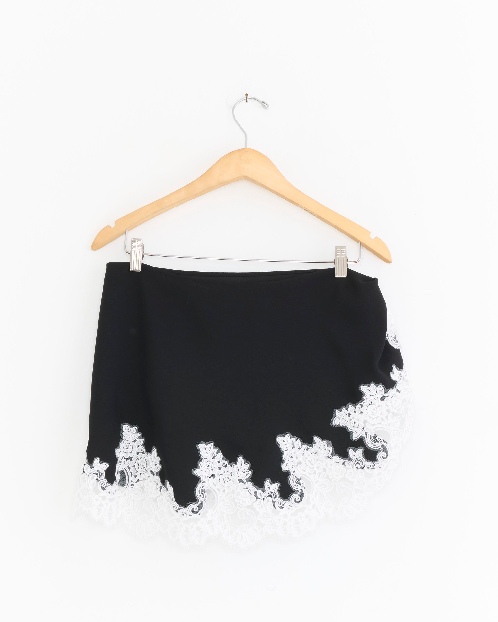 The Phoebe Wrap Skirt in Black by Beaufille features a scalloped hem and white embroidered trim, made from recycled polyester, shown on a wooden hanger against a plain white background.