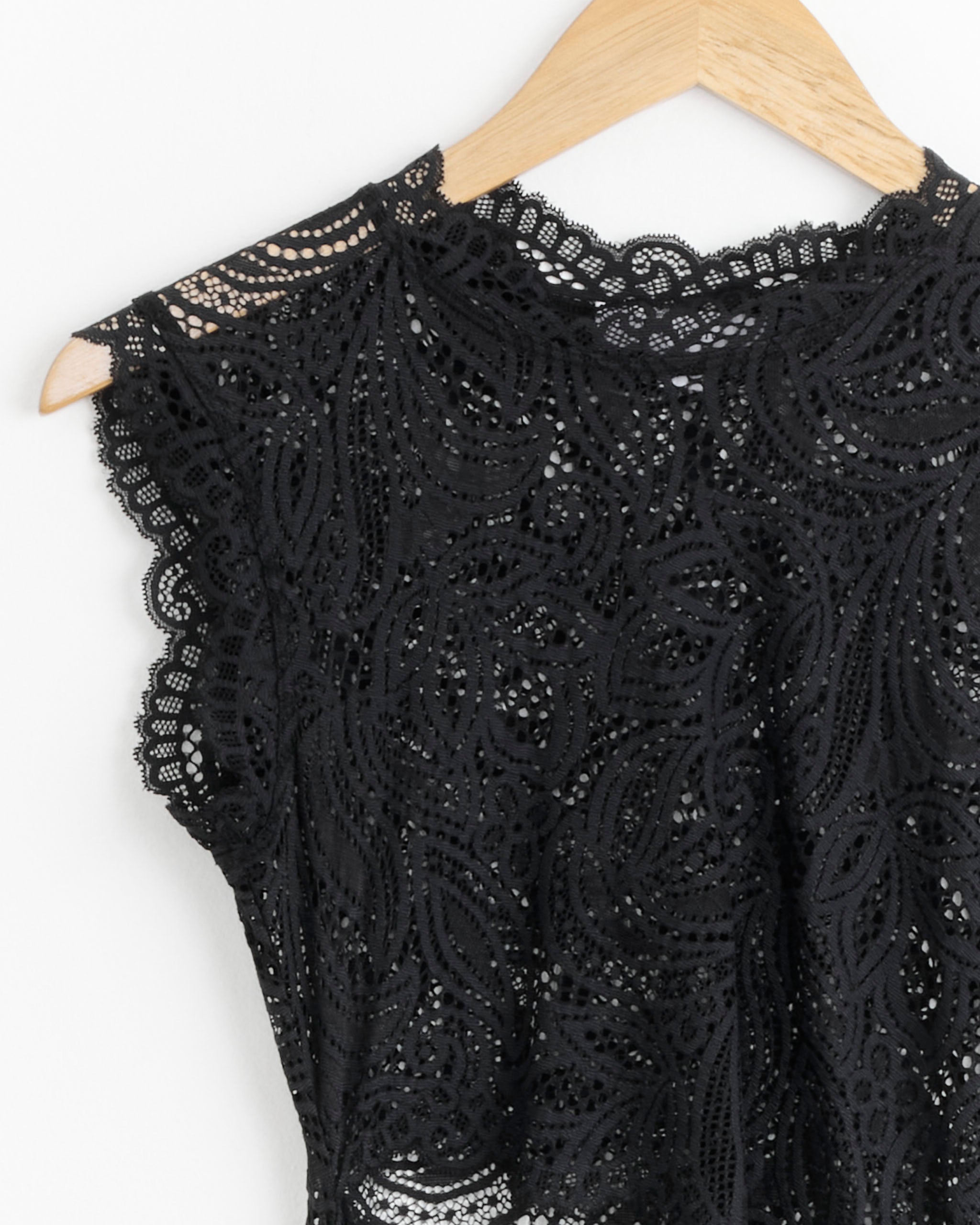 A close-up of the Silva Dress in Black by Beaufille, featuring intricate floral lace and delicate scalloped edges, displayed on a wooden hanger against a white background.
