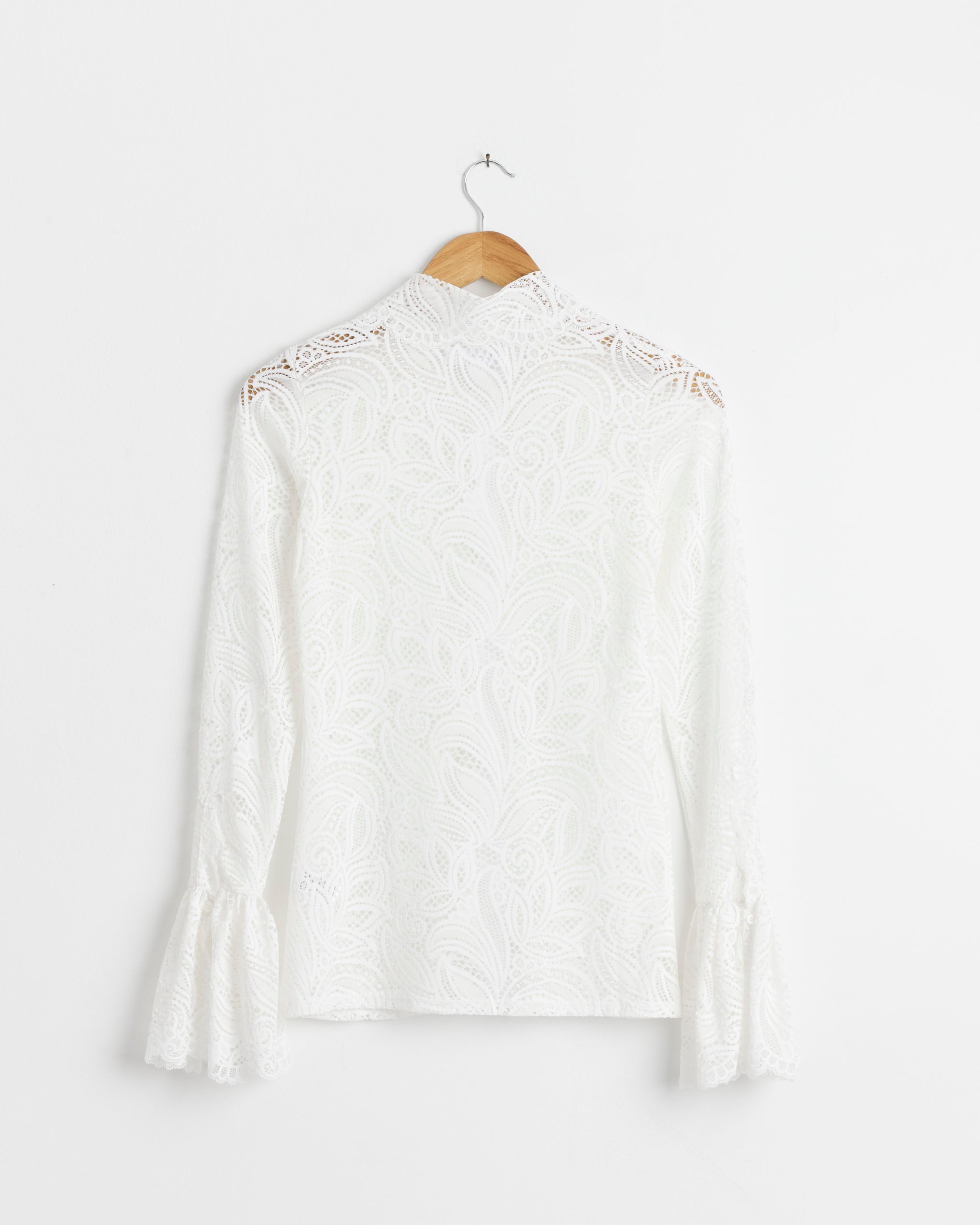 The Lucienne Blouse in White by Beaufille, a long-sleeve stretch lace top with a high neck, flared cuffs, and delicate leaf pattern, is displayed on a wooden hanger against a plain white wall.