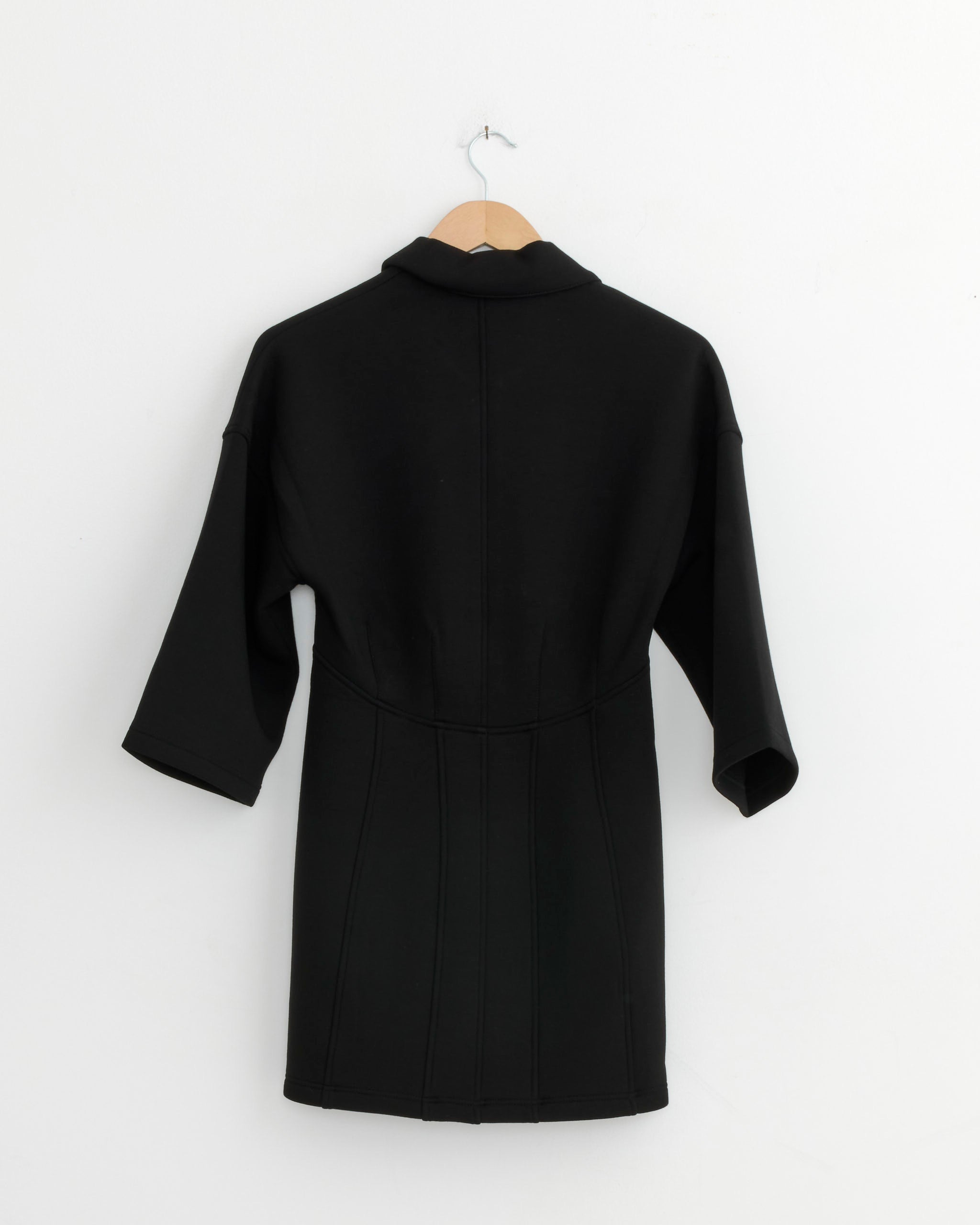 The Tibi Bonded Viscose Slim Tunic in Black, a knee-length, long-sleeve coat with a structured silhouette, is displayed from the back on a wooden hanger against a white wall.