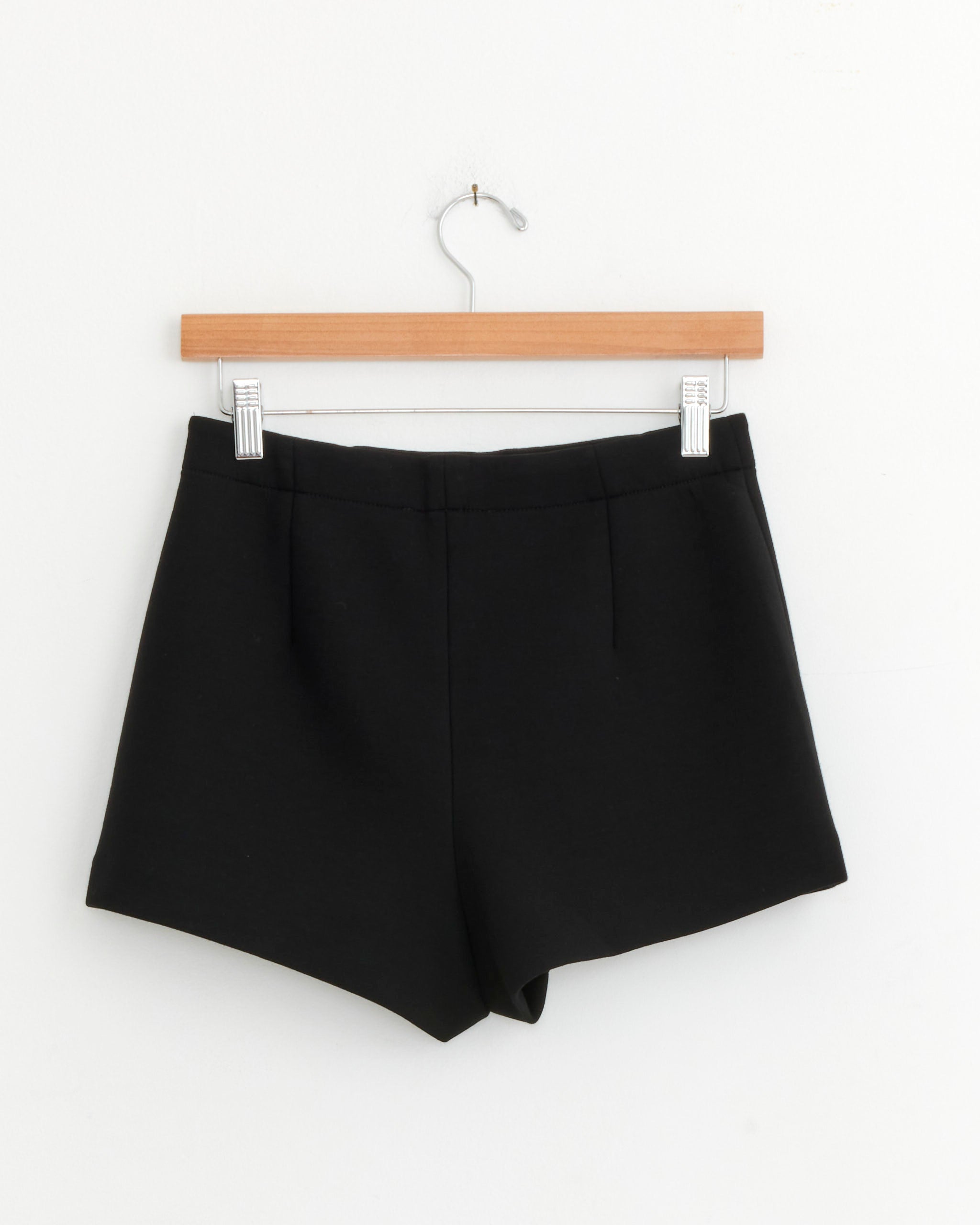 The Tibi Bonded Viscose Short in Black hangs on a wooden hanger against a white wall.