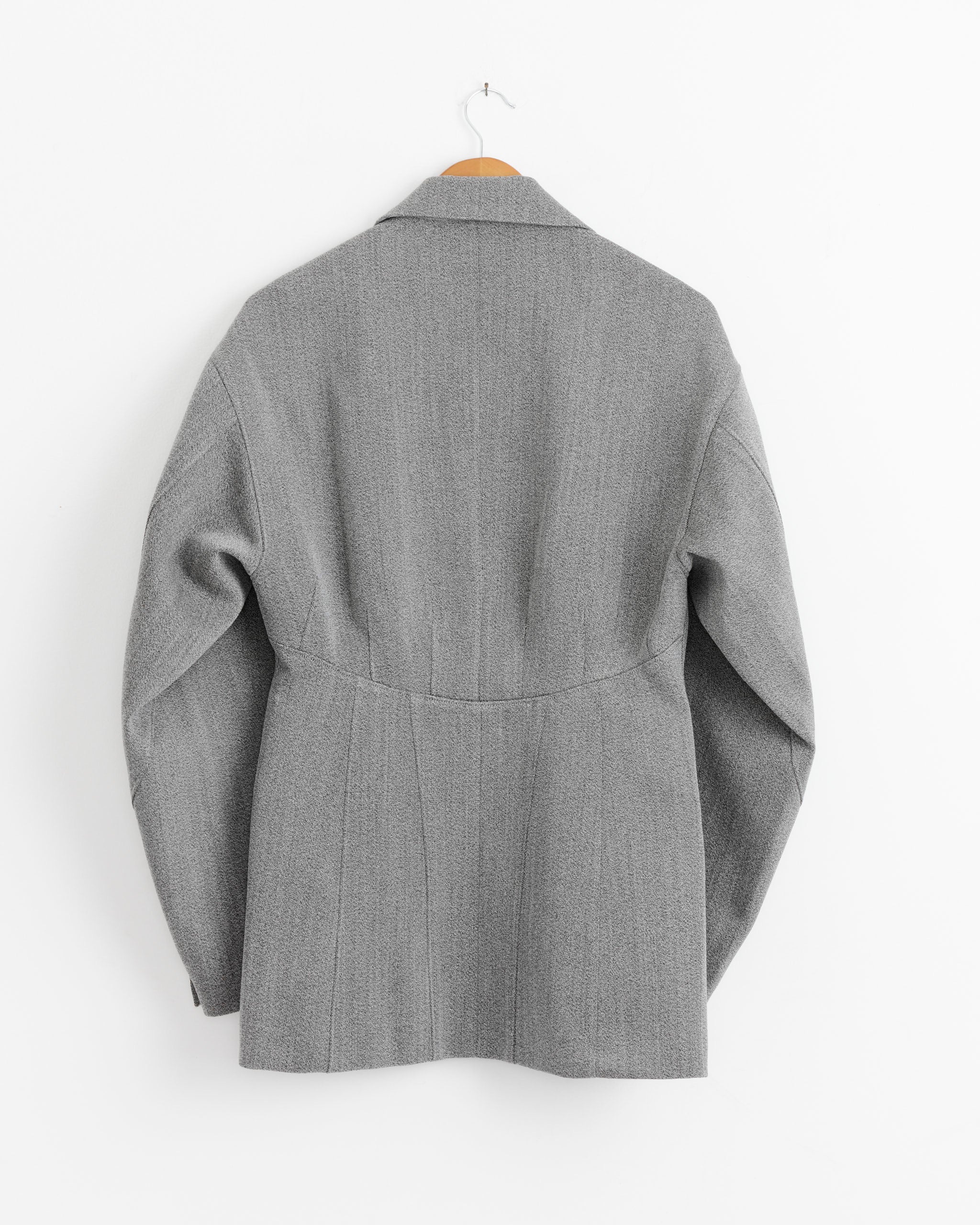 The Tibi Structured Wool Sculpted Blazer in Grey Multi hangs on a wooden hanger against a white wall, shown from the back to highlight its sculpted seams and precise tailoring.