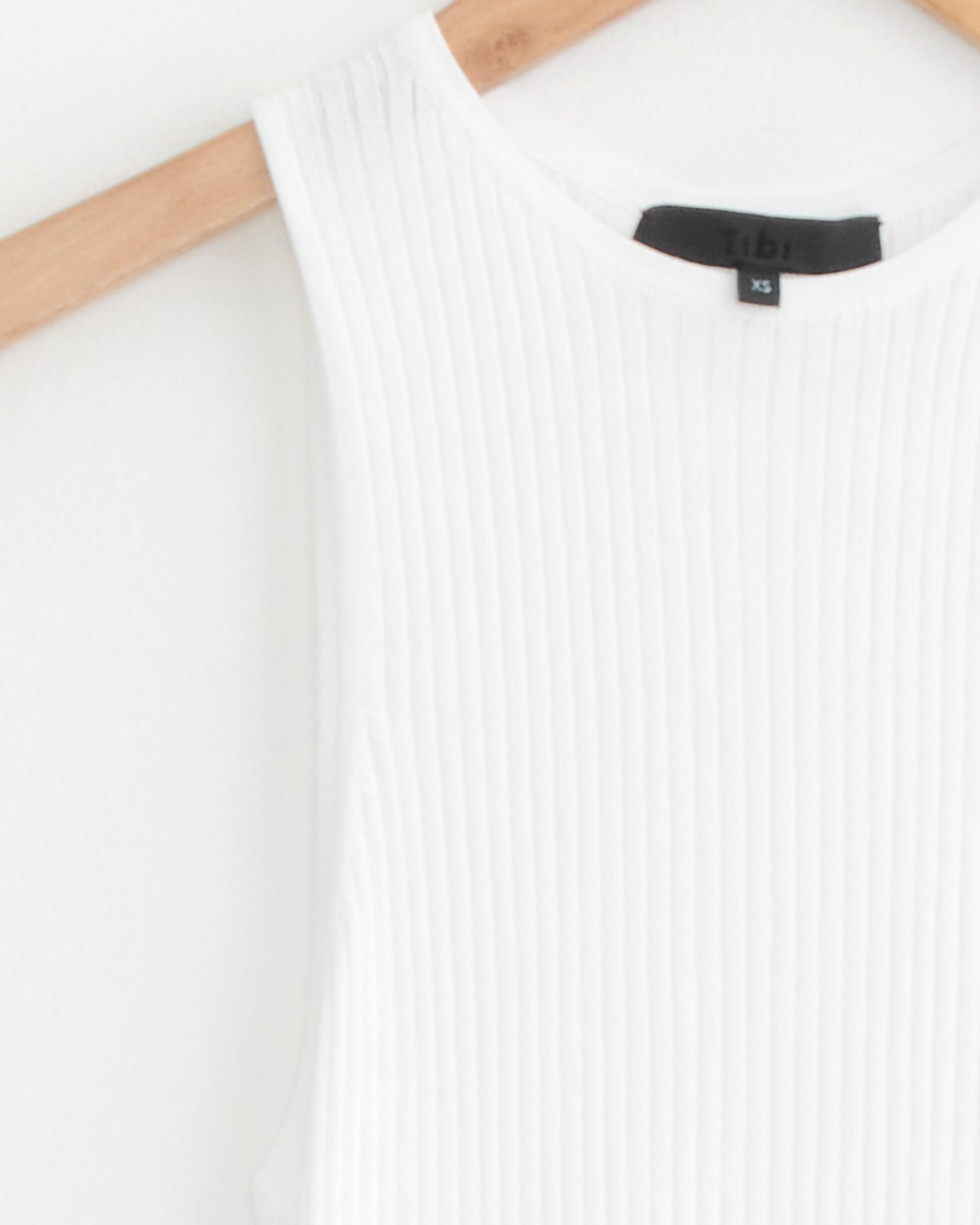 The image shows a close-up of the Tibi Ribbed Sweater Cut Out Tank in White, sleeveless with a round neckline and ribbed texture, displayed on a wooden hanger. A black label with "Tibi" and "XS" is visible against a plain white background.