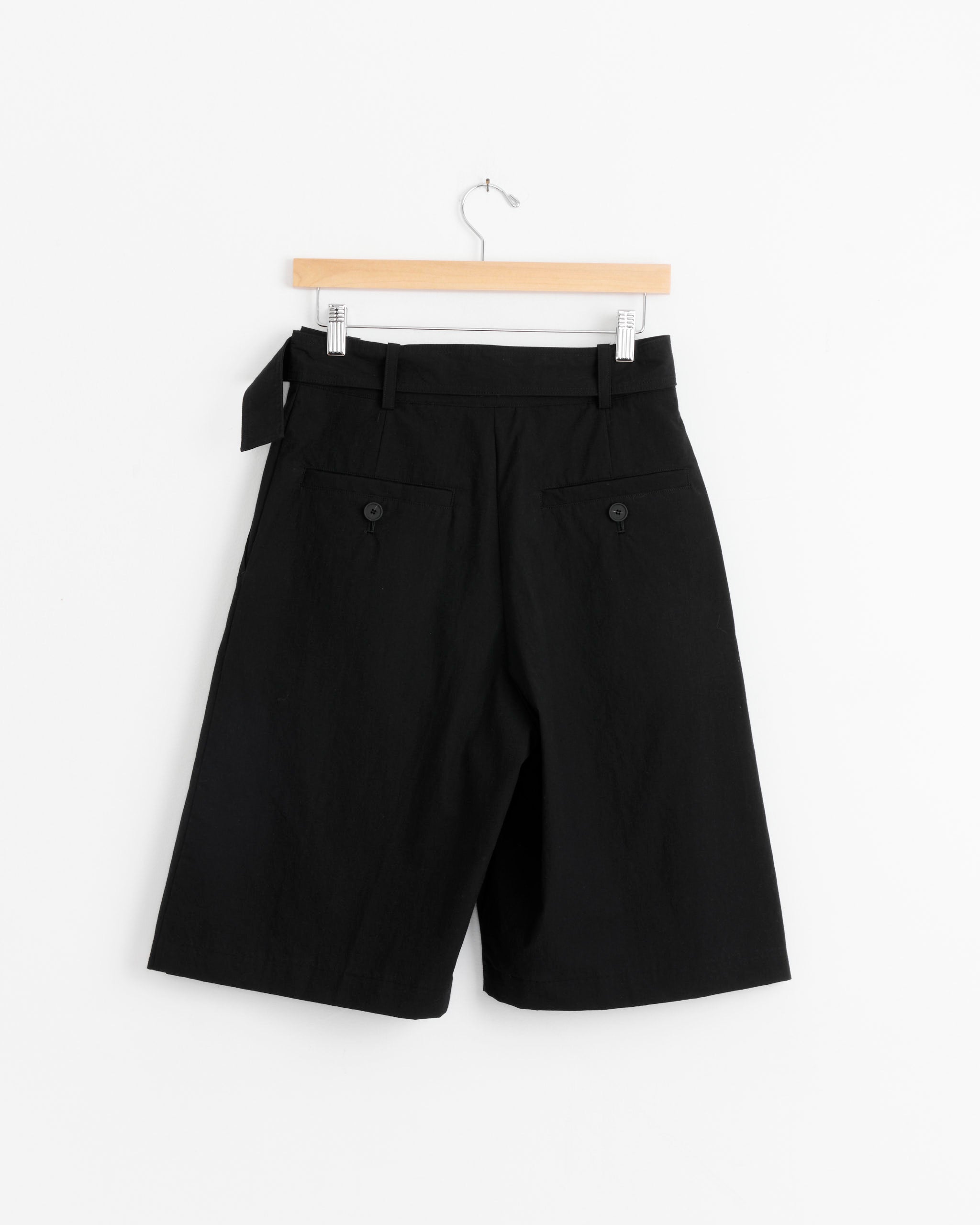The Creon Short in Black by Studio Nicholson features belt loops and back pockets, crafted from premium Japanese cotton, and is displayed on a wooden hanger against a plain white background.