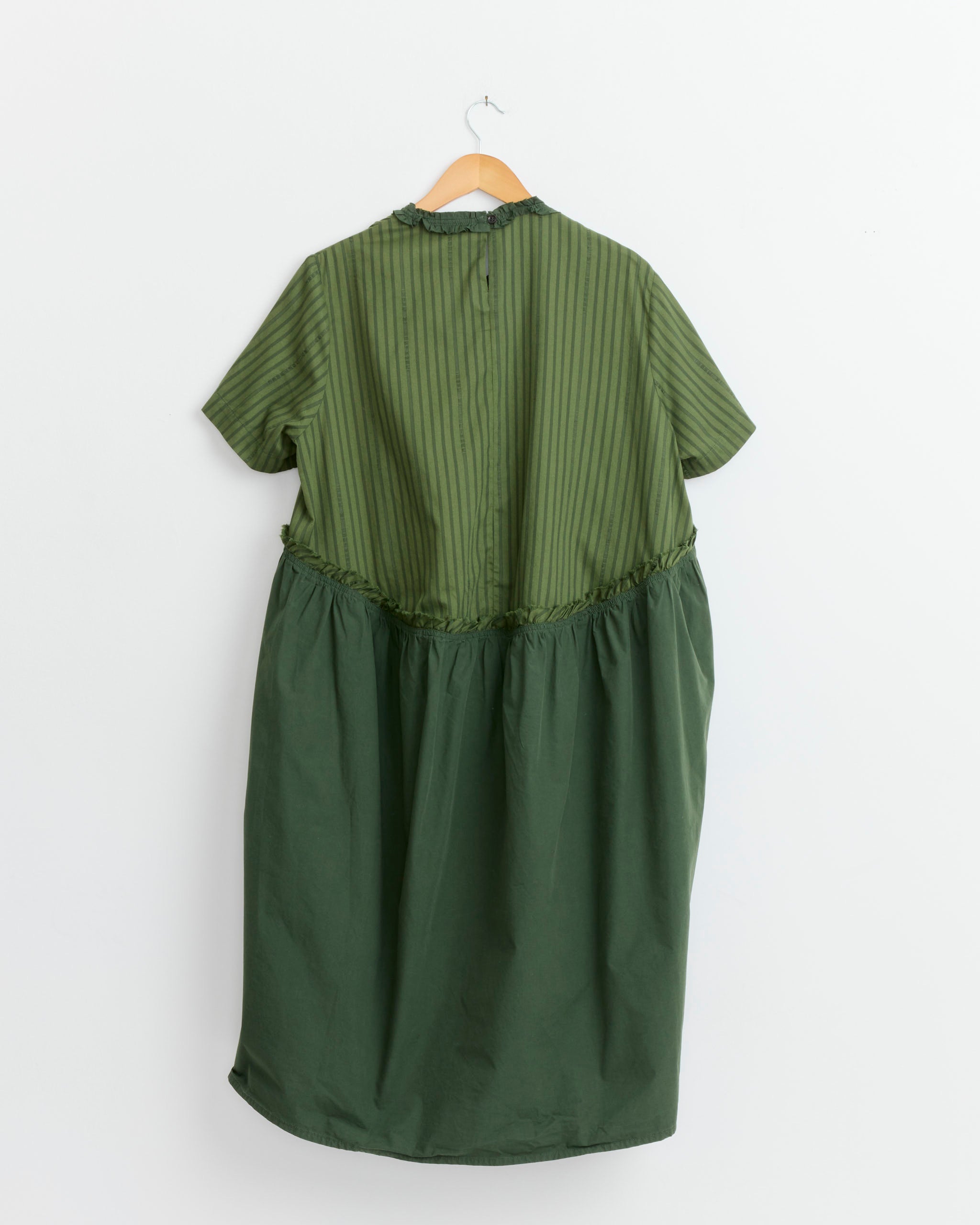 The Henrik Vibskov Bon Chalant Dress in Garden Green features organic cotton, short sleeves, a vertically striped top, and a solid green gathered skirt. It is displayed on a wooden hanger against a white background.