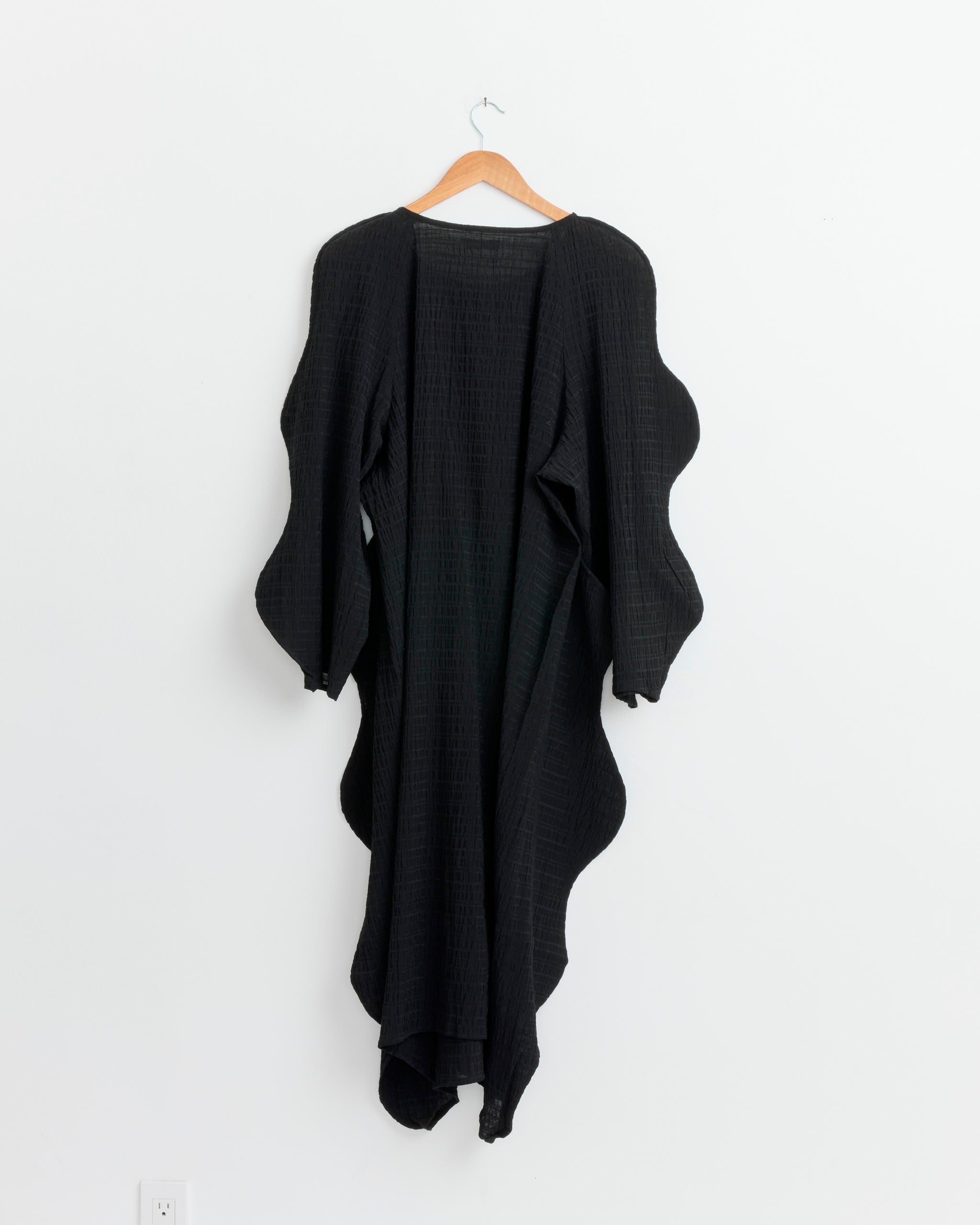 The Henrik Vibskov Octo Dress in Black, featuring textured checkered fabric and scalloped edges, hangs on a wooden hanger against a white wall.