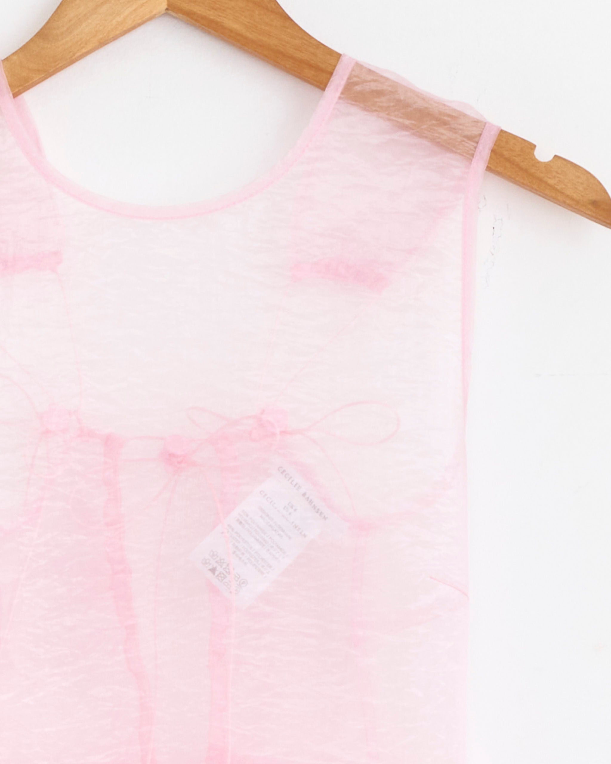The Cecilie Bahnsen Camilla Overlay Dress in Blush Pink Liquid Myrtia, crafted from delicate, translucent fabric, hangs on a wooden hanger against a white wall, revealing both the hanger and the inner seams.