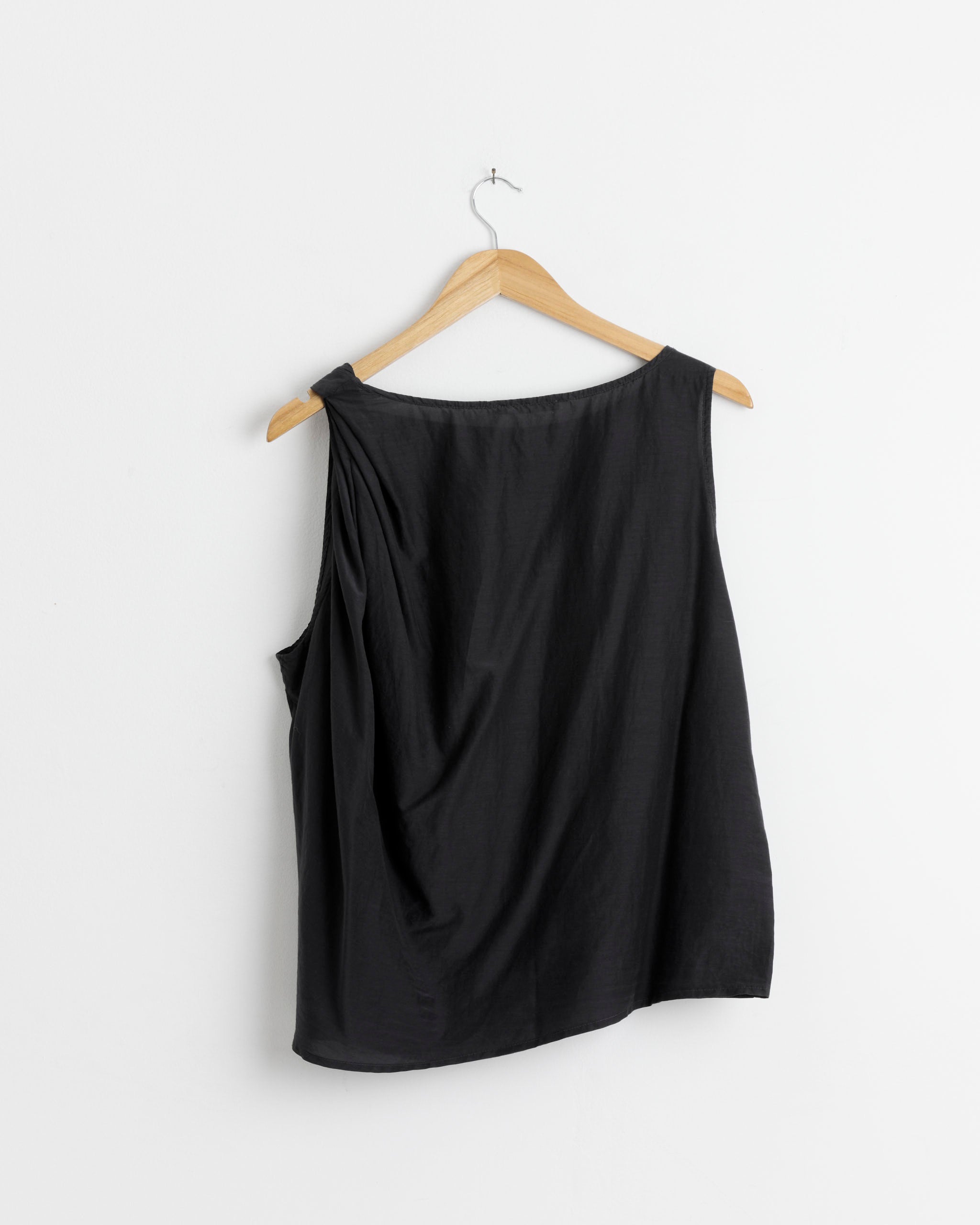The Black Crane Twisted Tank Top in Faded Black, made from a silk-cotton blend, hangs on a wooden hanger against a white wall. It features a loose, draped fit and wide neckline for effortless style.