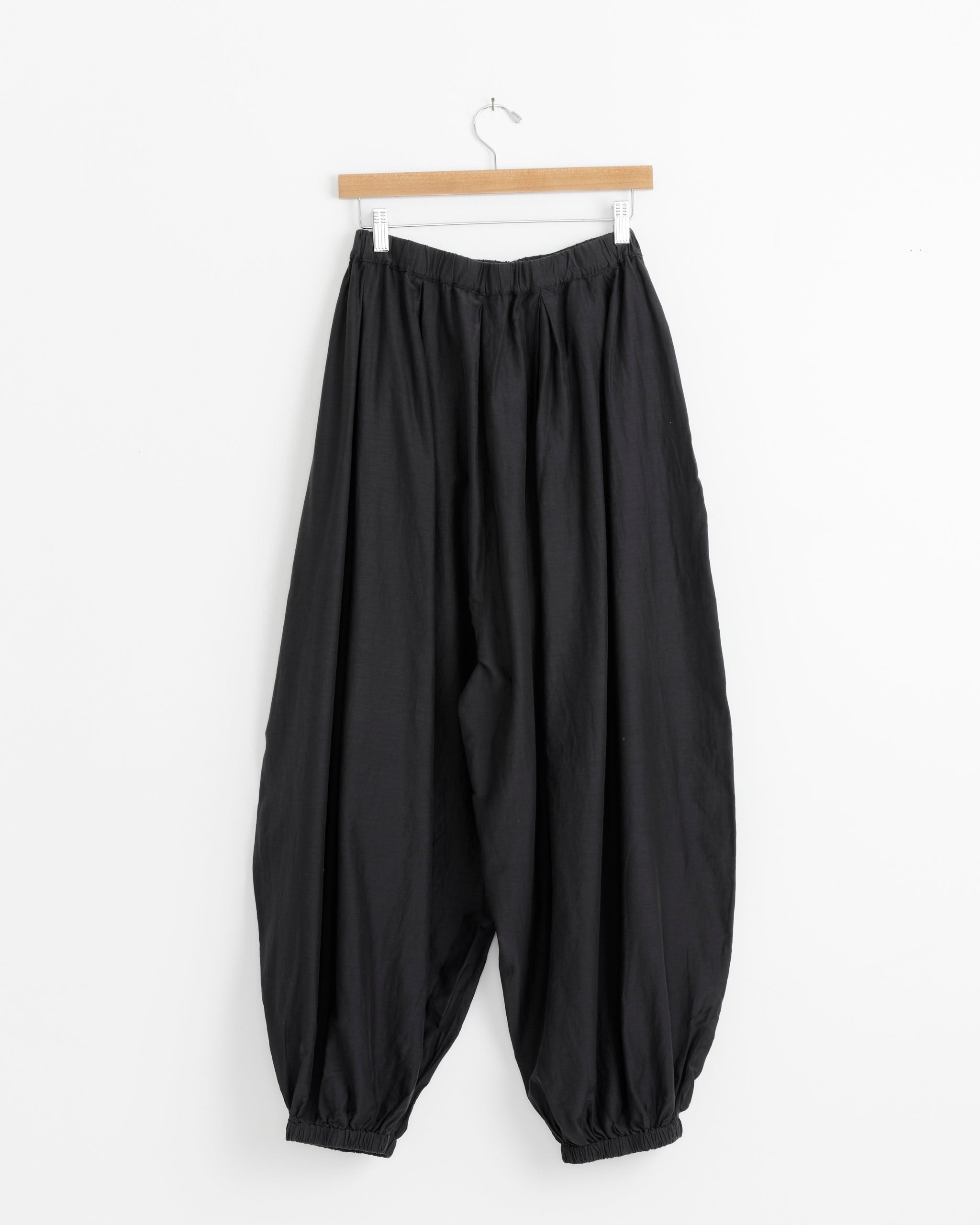 The Black Crane Akari Pant in Faded Black features a loose fit, elastic waistband, and balloon cuffs in soft silk cotton, shown hanging on a wooden hanger against a plain white background.