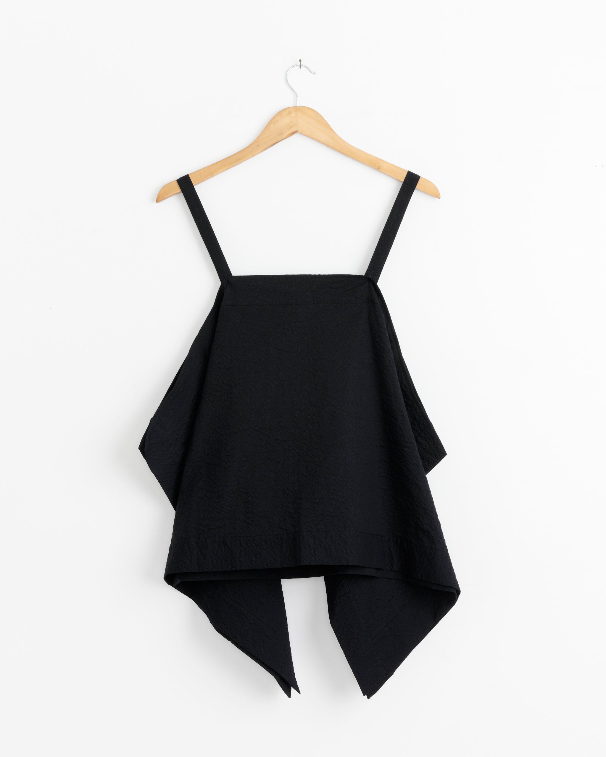 The Black Crane Paper Top in Midnight is a sleeveless black top made from Japanese textured cotton, with wide straps and an asymmetrical handkerchief hem, shown on a wooden hanger against a white background.