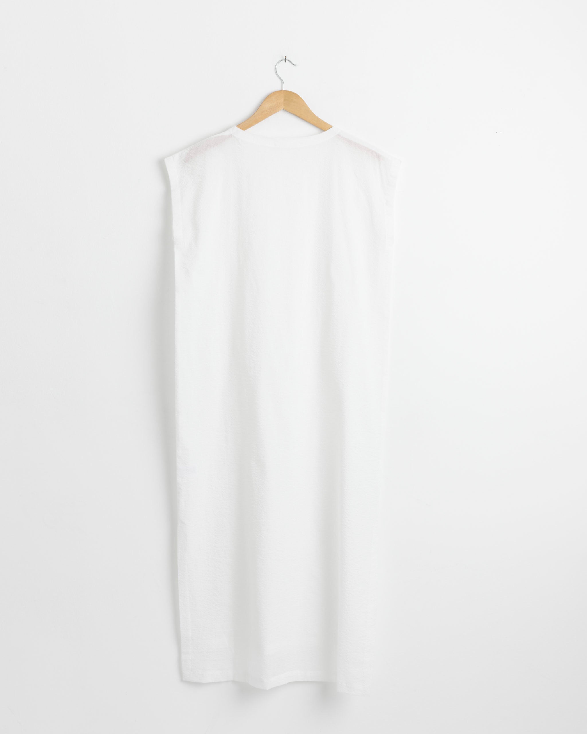 The No Sleeve Dress in White by Black Crane, an ankle-length textured cotton dress, is displayed from the back on a wooden hanger against a plain white wall.