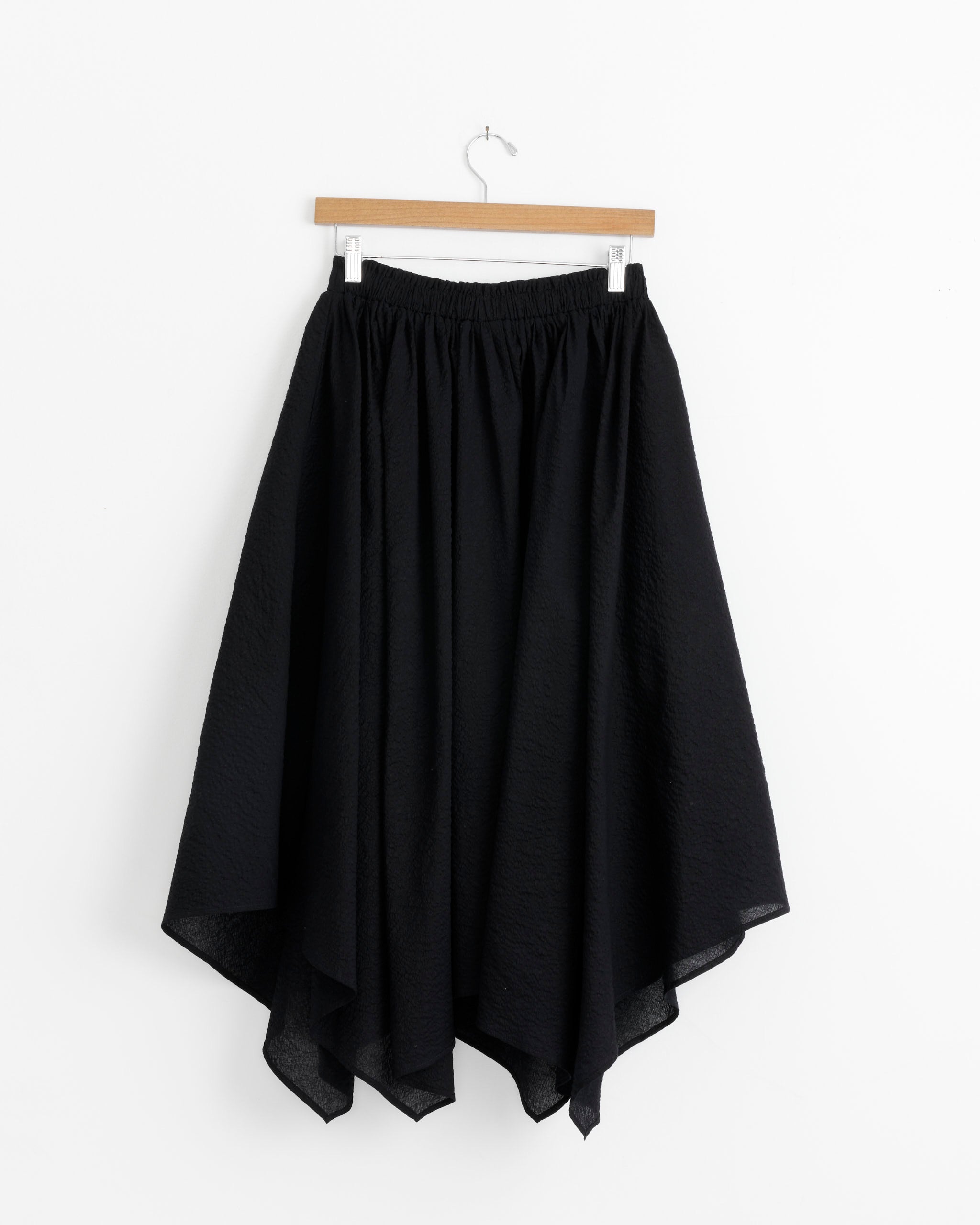 The Black Crane Paper Skirt in Midnight, a flowy, handkerchief-hem skirt made from Japanese textured cotton with an elastic waistband, is displayed on a wooden hanger against a white background.