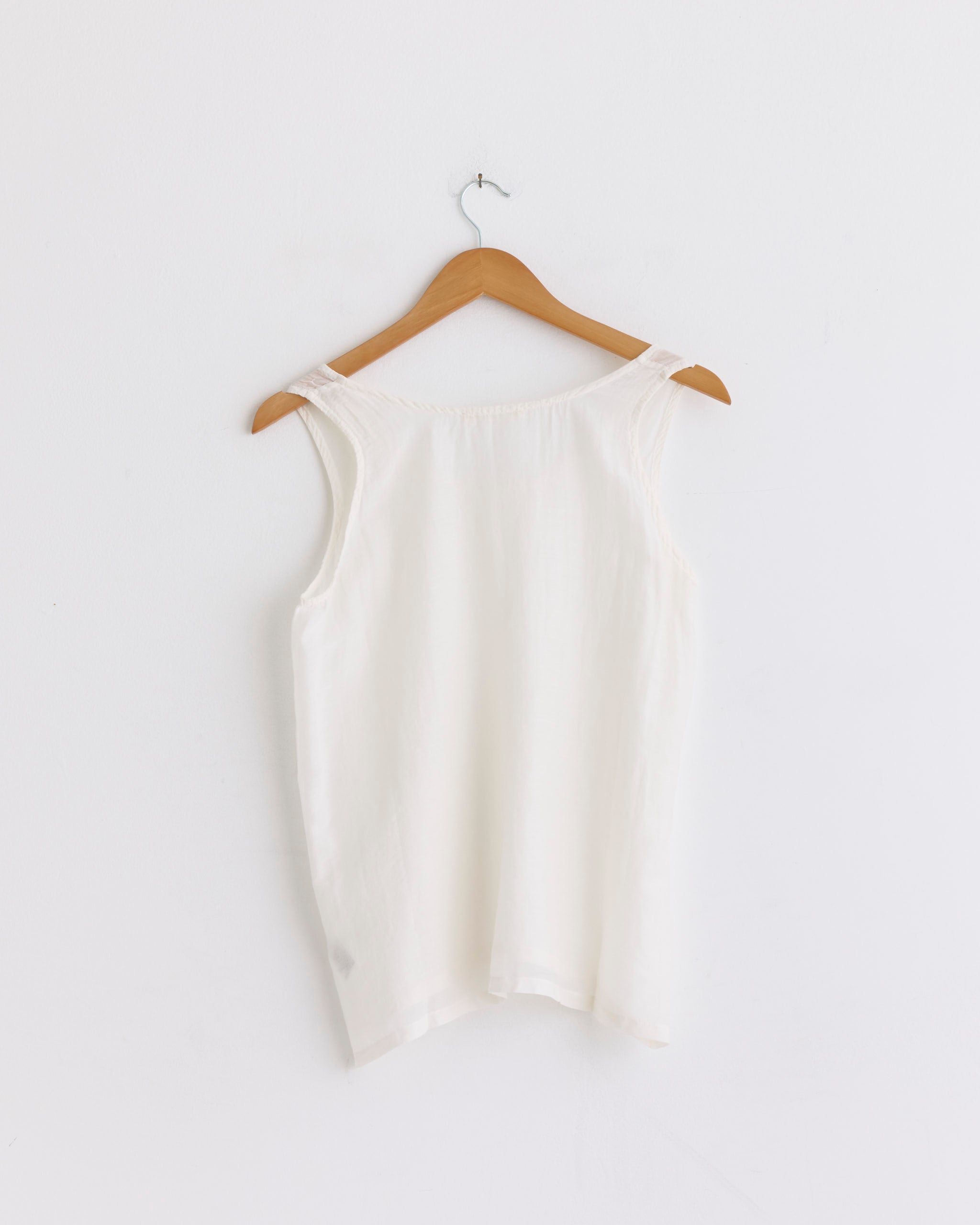 The Loose Tank Top in White by Black Crane hangs on a wooden hanger against a plain white wall, showcasing its minimal and elegant silhouette.