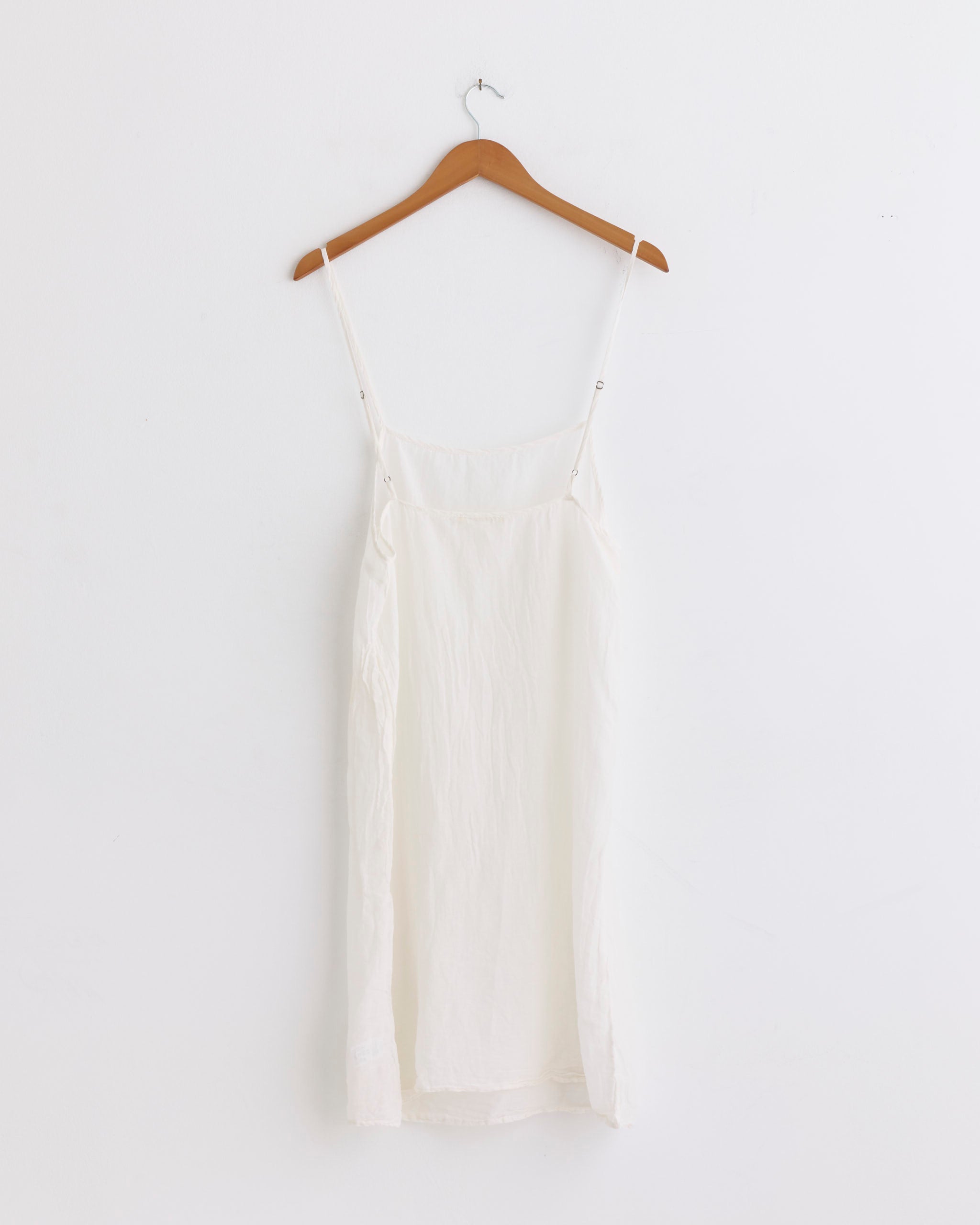 The Black Crane Cami Dress in White, made from silk cotton, hangs on a wooden hanger against a white wall. This sleeveless dress features thin straps and a relaxed silhouette.