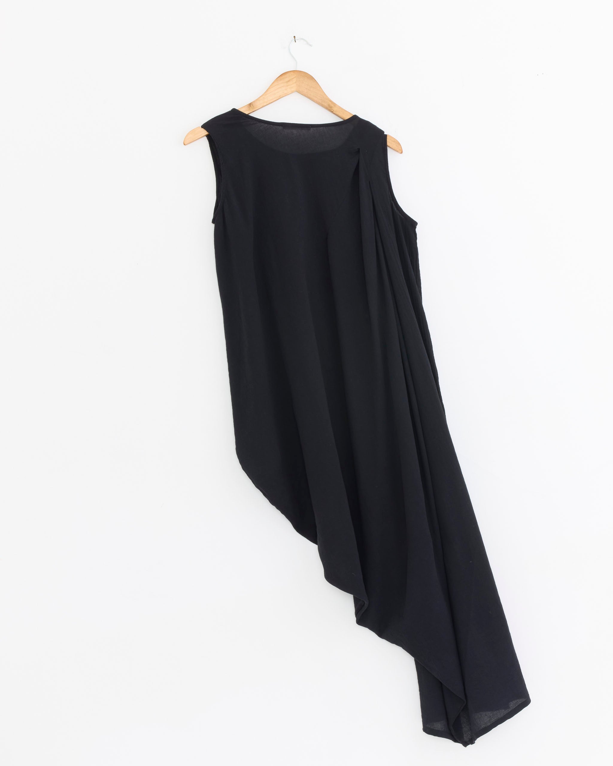 The Black Crane Greek Column Dress in Sumi Black, made of cotton gauze with a sleeveless design and asymmetrical draped hem, hangs on a wooden hanger against a white background.