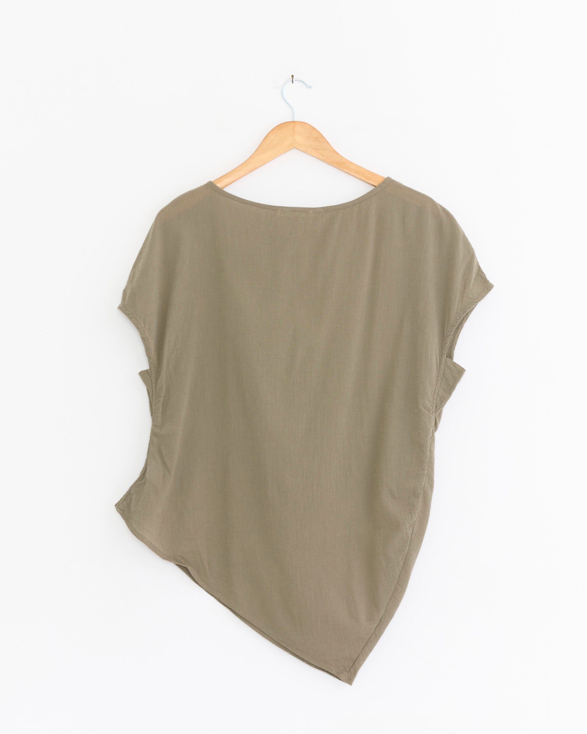The Black Crane Asymmetric Top in Cement, made from textured cotton gauze, hangs loose with a slanted hemline and short sleeves. Displayed on a wooden hanger against a white background. Made in USA.