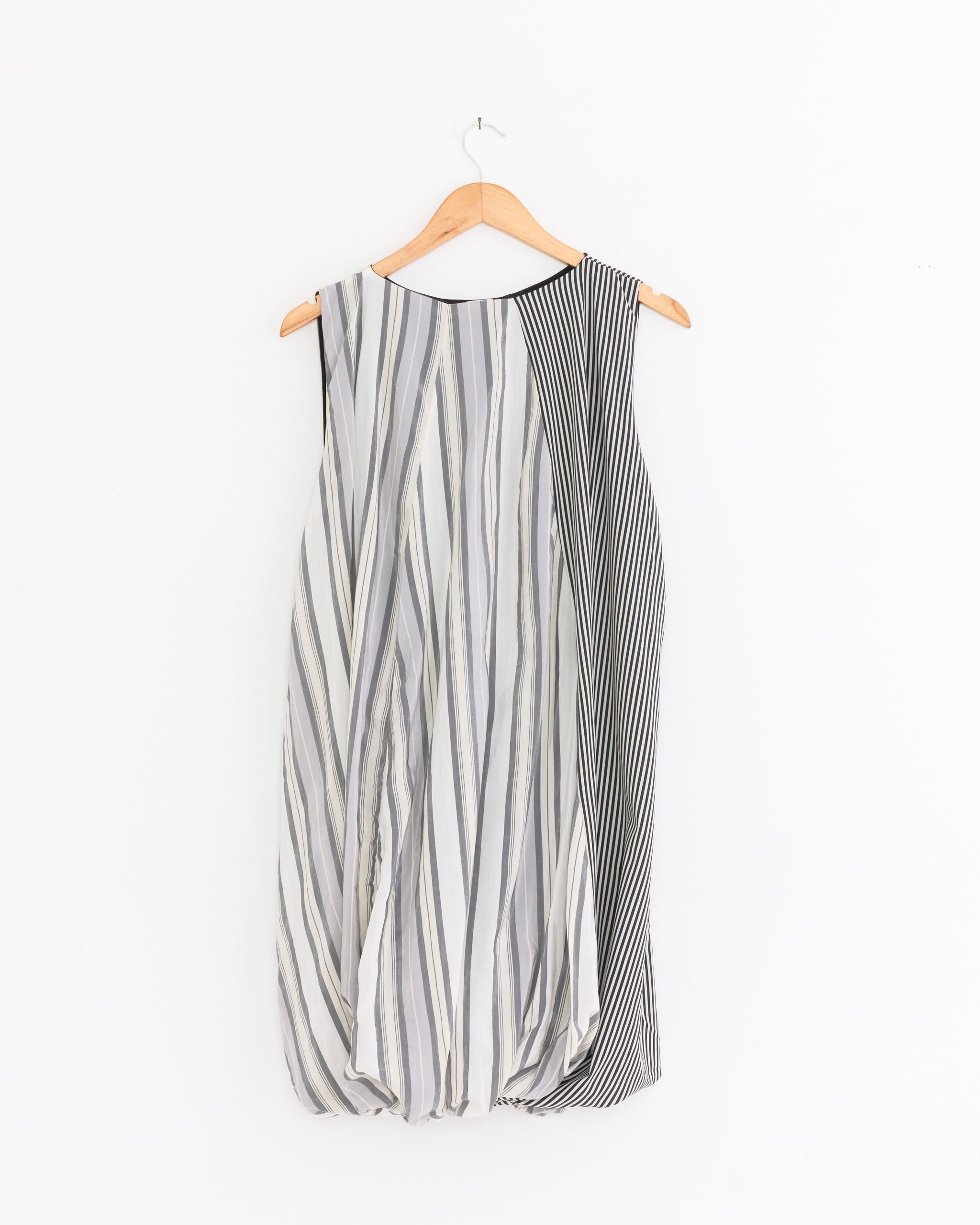 The Baserange Bella Dress in Grey Stripe, shown on a wooden hanger against a white background, features vertical and diagonal black, white, and gray stripes for an asymmetrical, modern look.