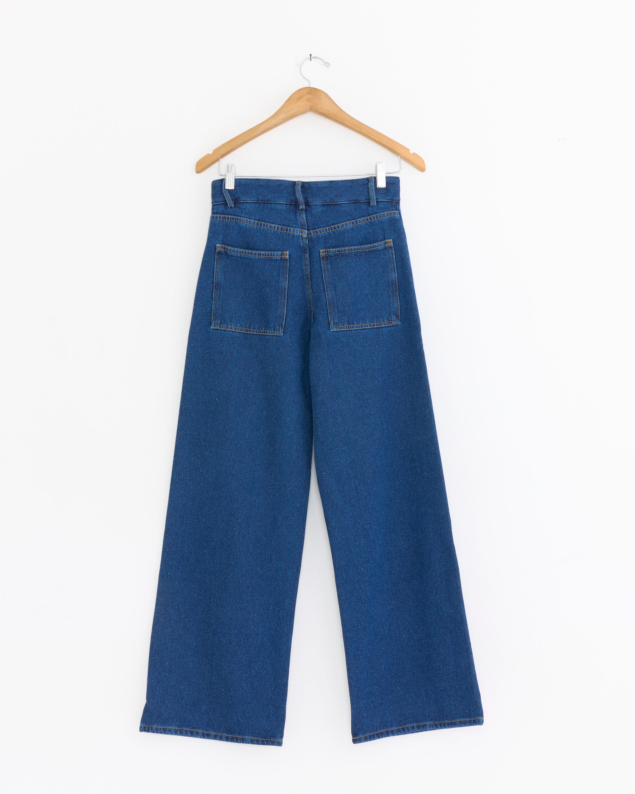 The Grass Pant in Indigo Blue Wash by Baserange, featuring a high waist and visible back pockets, hangs on a wooden hanger against a white background with its wide-leg denim silhouette shown from the back.
