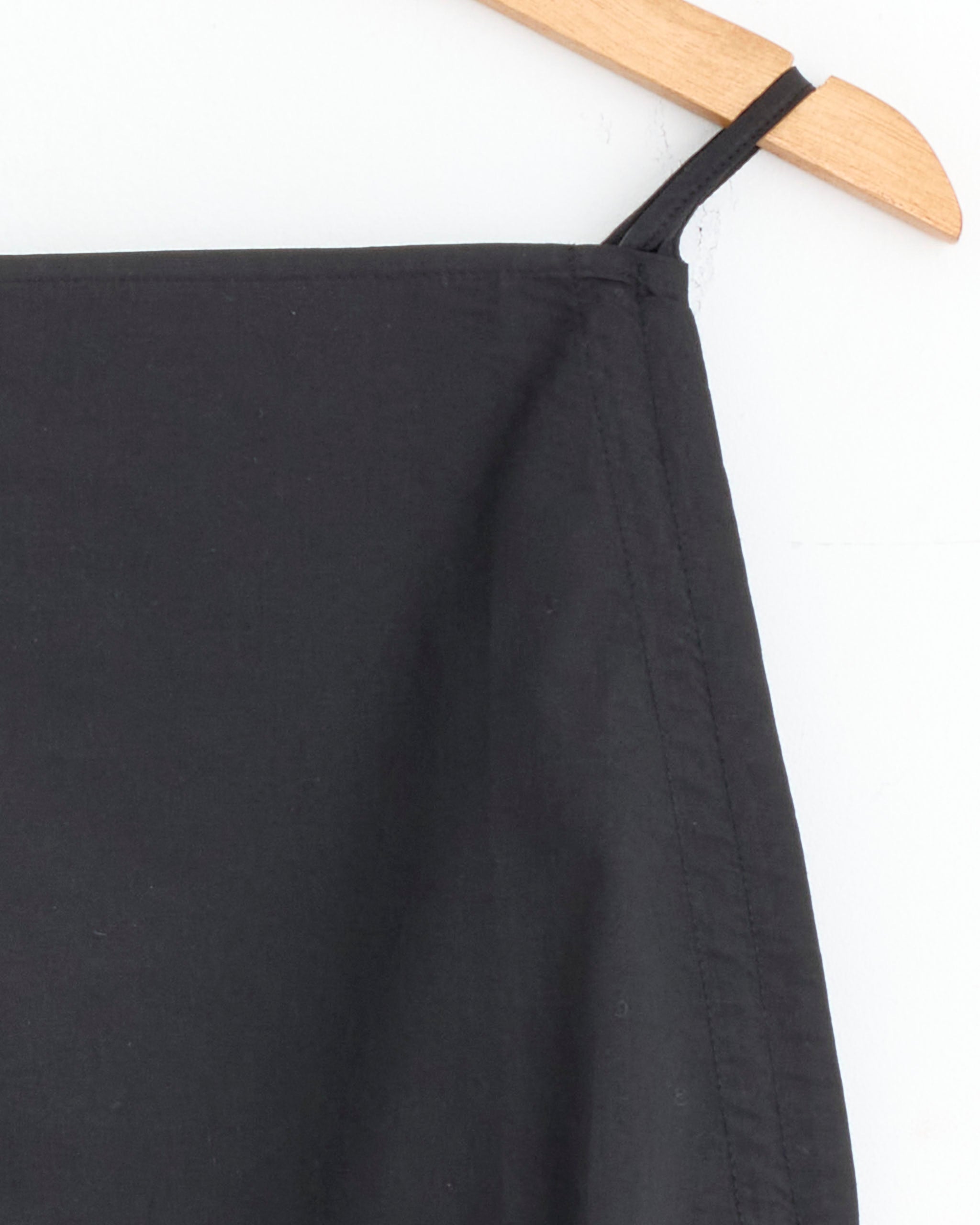 Close-up of the top corner of the Baserange Pictorial Top in Black with adjustable straps, shown on a wooden hanger against a white background. Only part of the garment and hanger is visible.