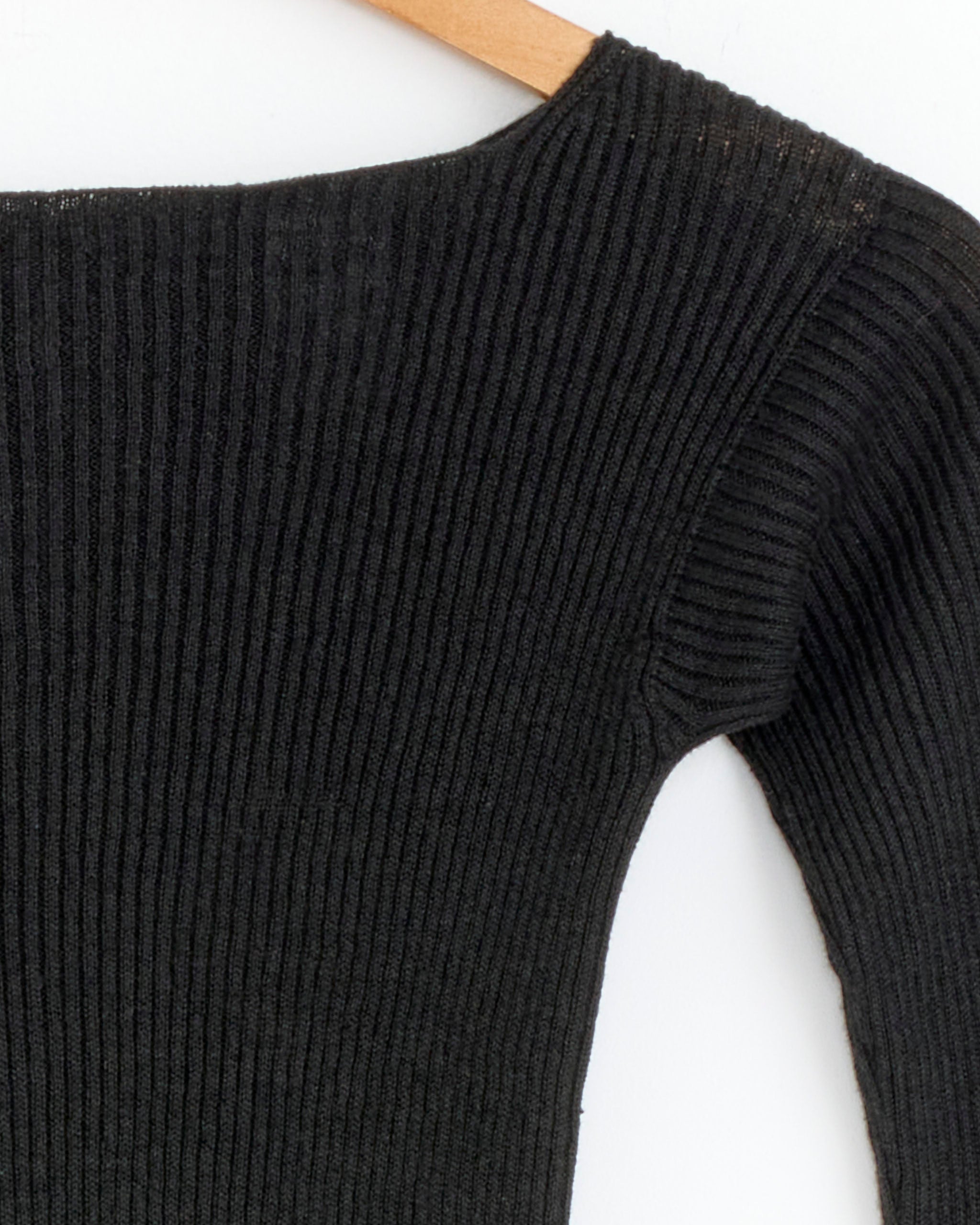 Close-up of the Baserange Oder Top in Black, a ribbed long sleeve style, hanging on a wooden hanger against a white background. Only the upper part and one sleeve are shown.