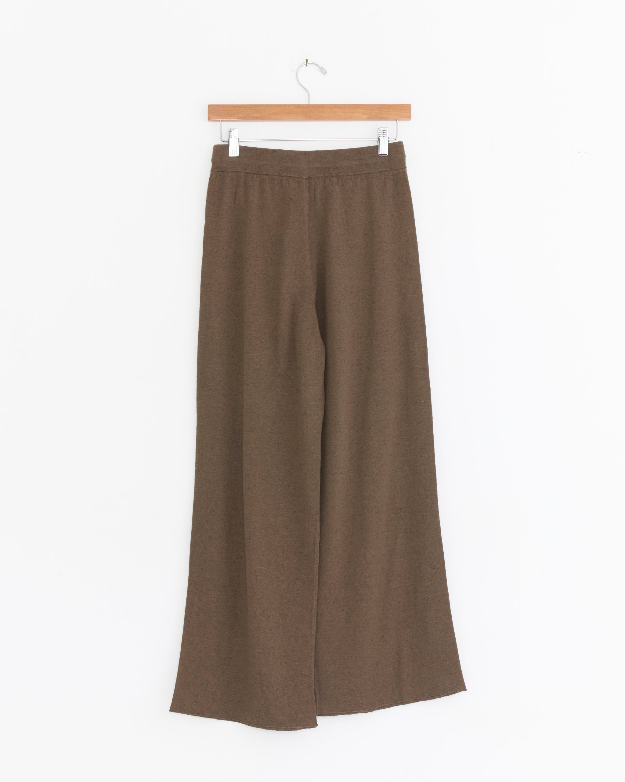 The Shaina Mote Chill Pant in Bark hangs on a wooden hanger against a white background. These wide-leg, high-waist pants are crafted from soft cotton linen with an elastic waistband and relaxed, loose fit.
