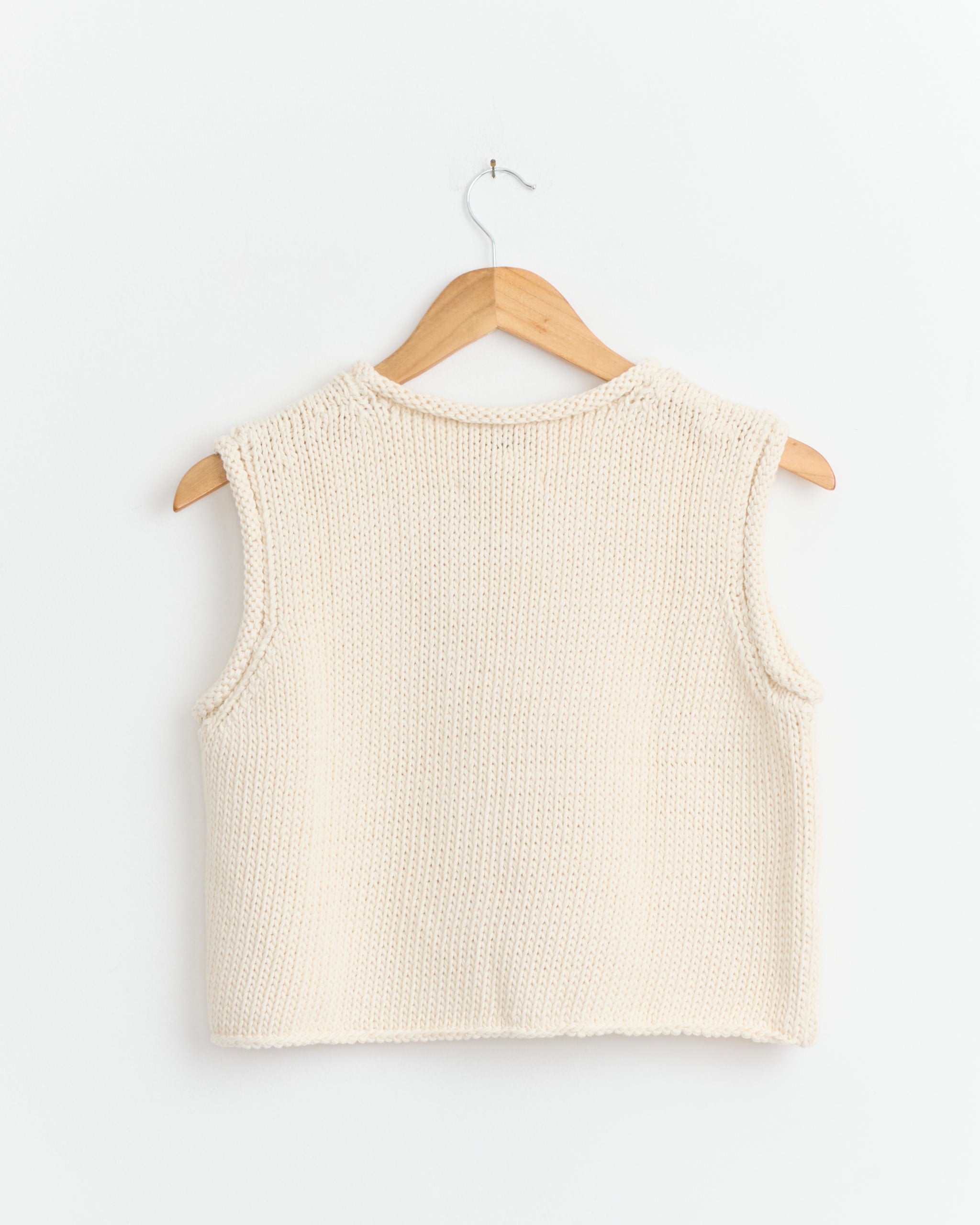 The Shaina Mote Brisa Top in Natural—a sleeveless, cropped shell—hangs on a wooden hanger against a white wall, offering effortless and timeless style.