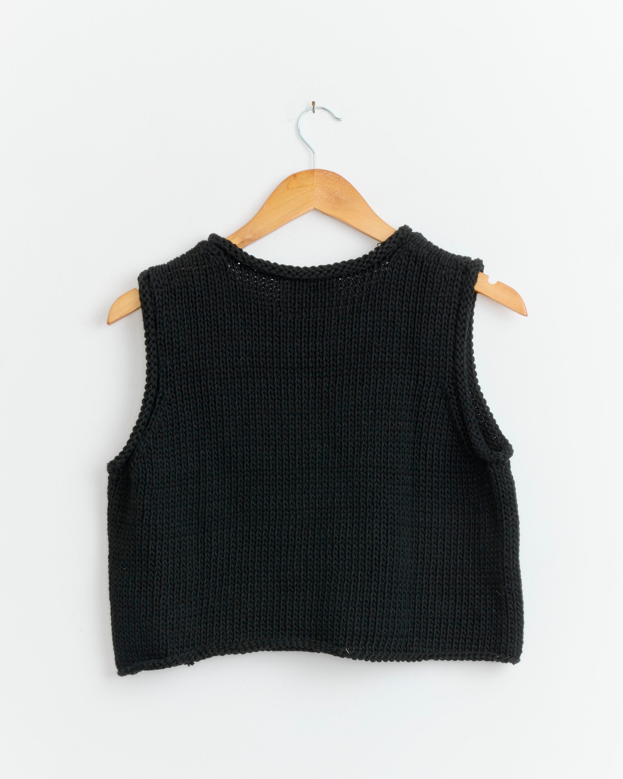 The Shaina Mote Brisa Top in Onyx, a sleeveless handknit piece made from organic cotton, hangs on a wooden hanger against a plain white background.