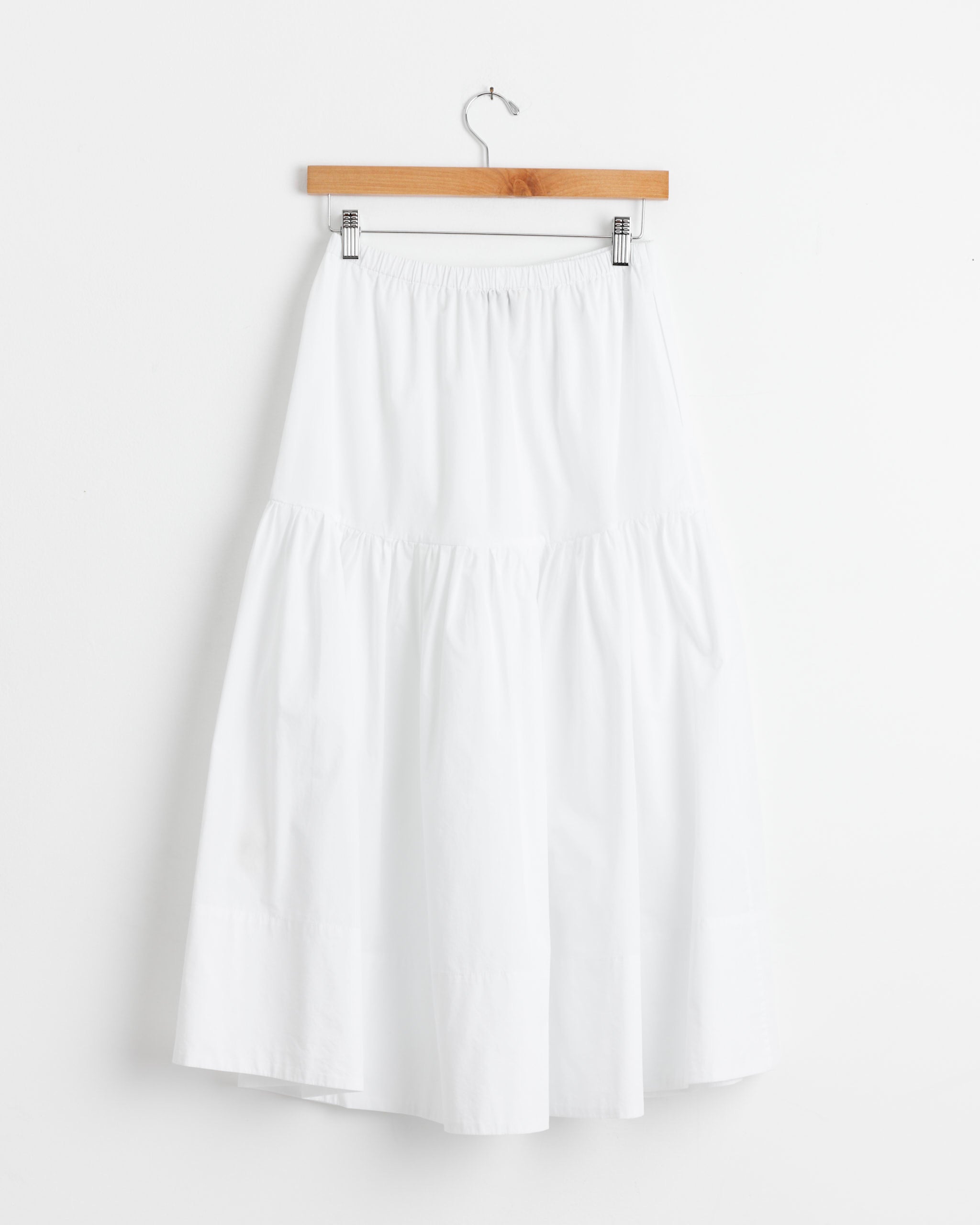 The Shaina Mote Gilot Skirt in Salt, a white, knee-length organic cotton skirt with an elastic waistband, hangs on a wooden hanger against a plain white wall.