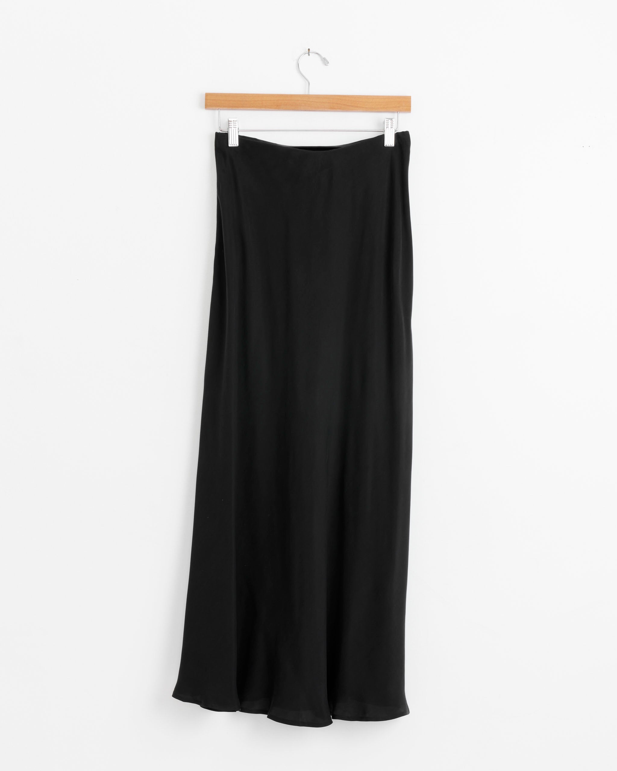 The Shaina Mote Bias Skirt in Onyx, long and silky, hangs on a wooden hanger against a plain white background.