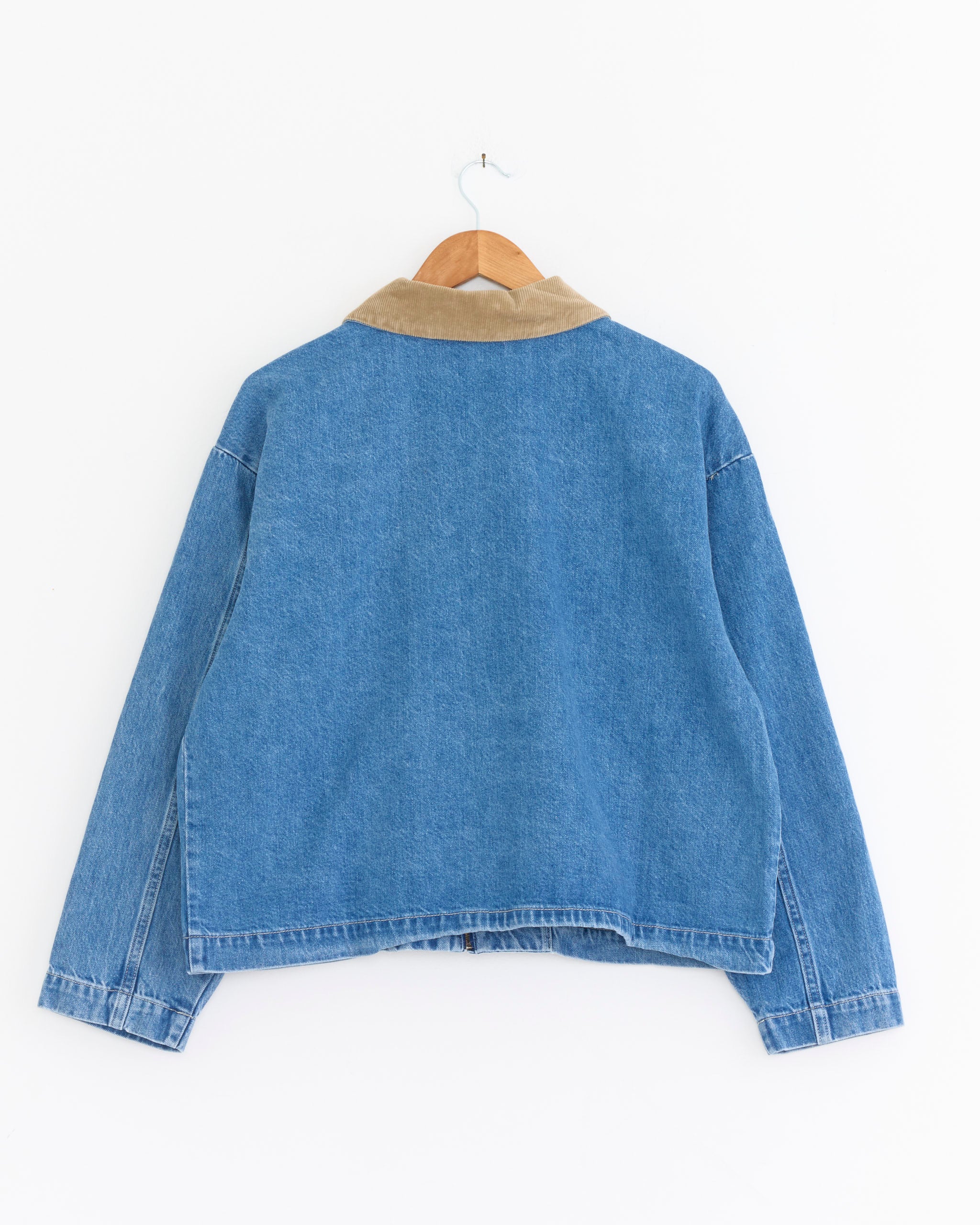The Shaina Mote Studio Coat in Medium Blue features a tan corduroy collar. Shown from the back on a wooden hanger against a white background, this jacket is proudly made in the USA.