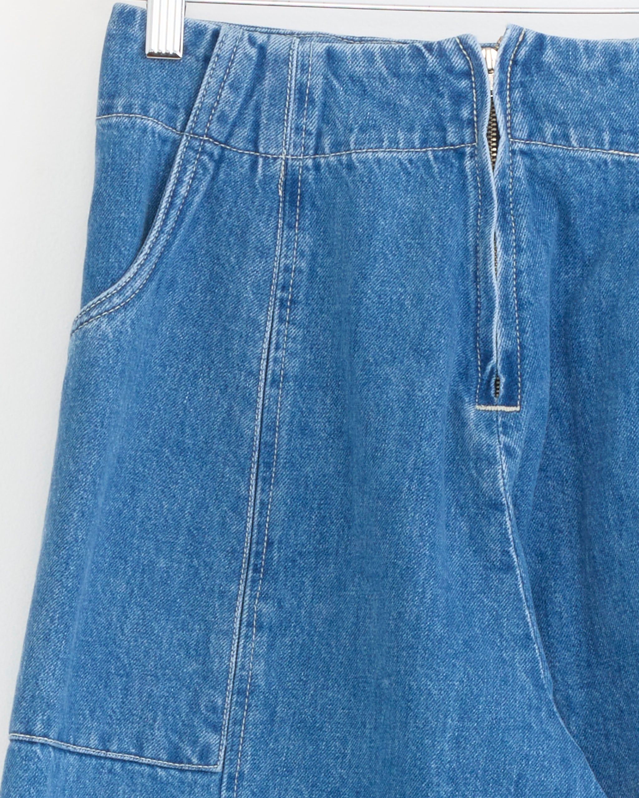 Close-up of the Sal Pant in Medium Blue by Shaina Mote, crafted from premium Italian denim with a front zipper and side pocket, hanging on a hanger against a light background.