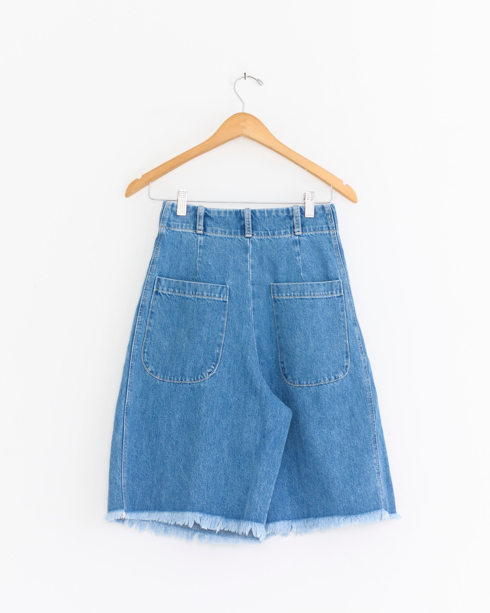The Emil Short in Vintage Indigo by Shaina Mote, crafted from BCI cotton with frayed hems and two large back pockets, is displayed on a wooden hanger against a plain white background.