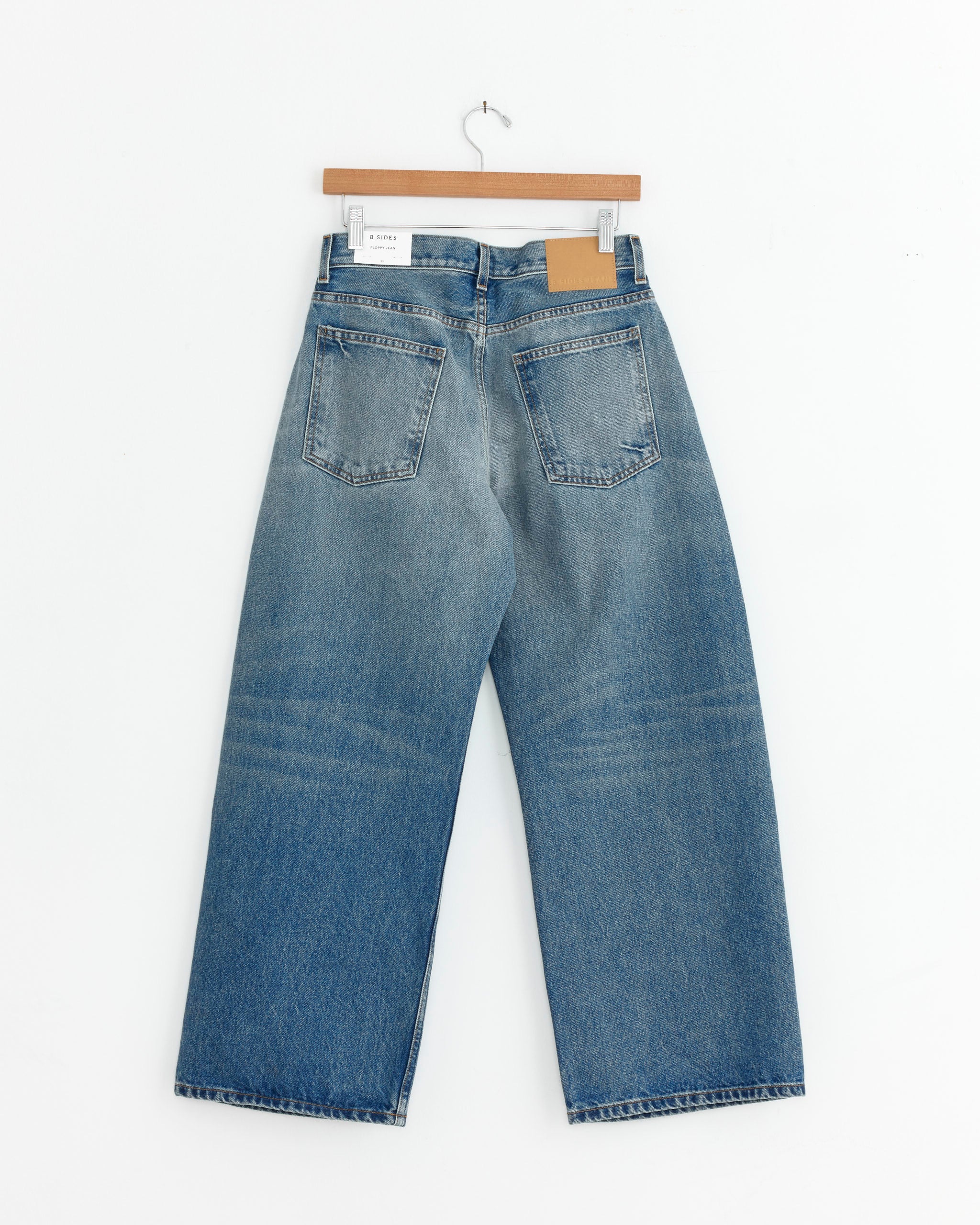 The B Sides Floppy Jean in Charley Wash, featuring an indigo hue, wide-leg cut, two back pockets, and a brown waistband patch, is displayed on a wooden hanger against a white background—timeless style made in the USA.
